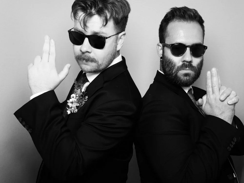 Two men in black suits and sunglasses posing back-to-back with serious expressions, one with a beard and the other with a mustache, against a plain background.