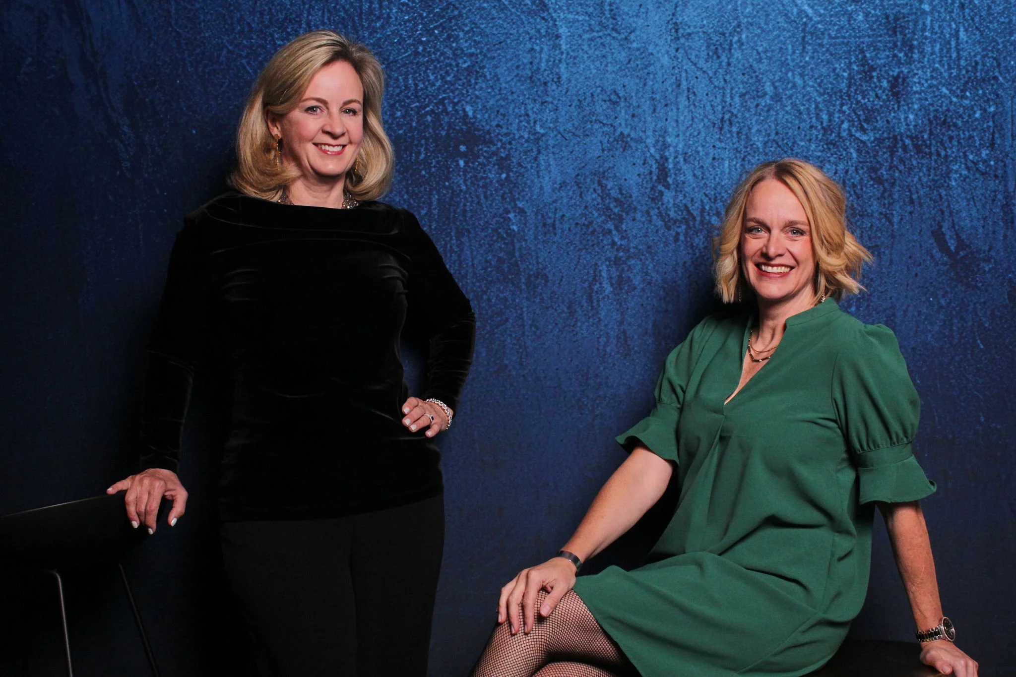 Two women posing against a textured blue wall. One woman stands, wearing a black long-sleeve top and black pants, with blonde hair styled in loose waves. The other woman is seated on a black chair, wearing a green dress with puff sleeves, fishnet sto