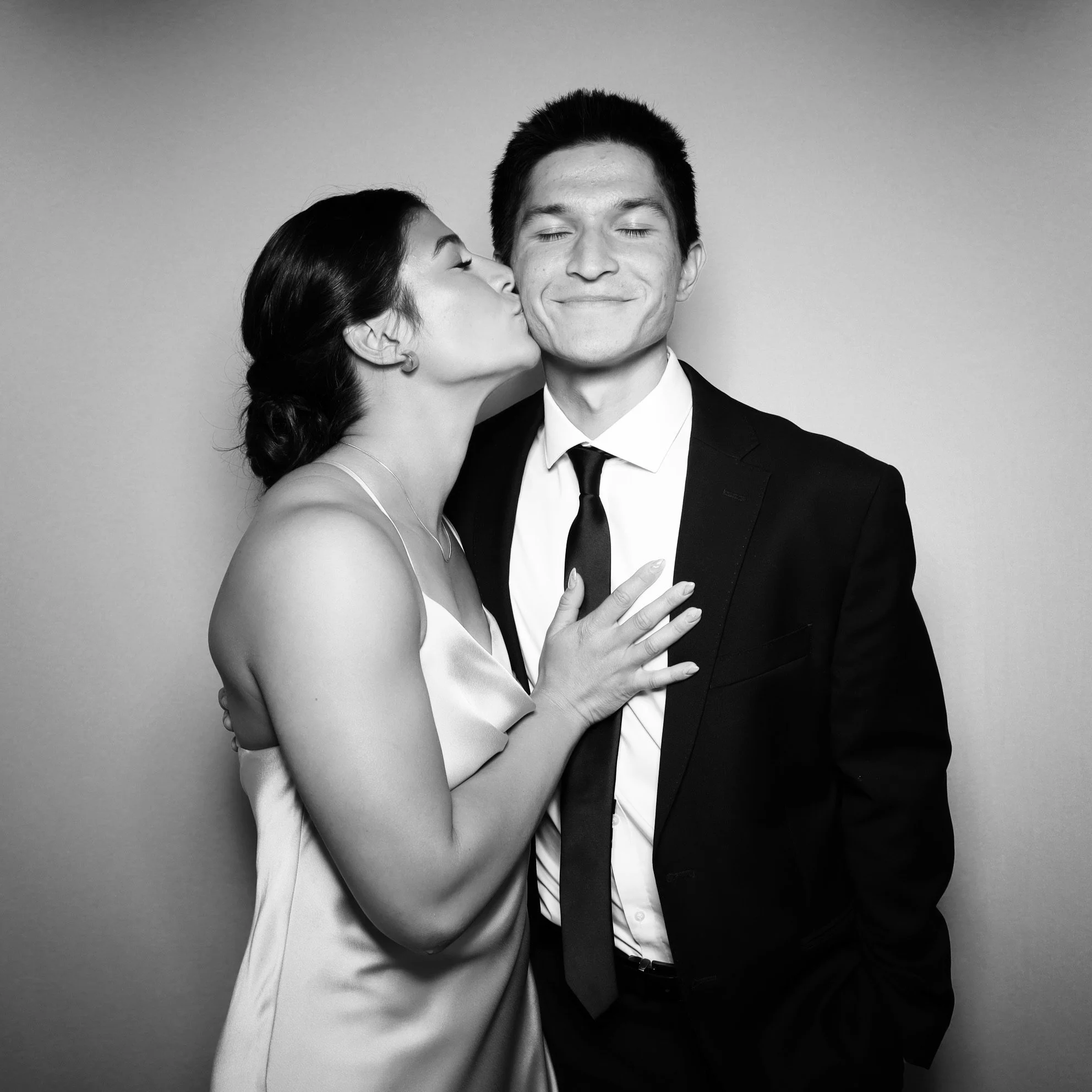 A woman in a satin dress kisses a smiling man in a tuxedo on the cheek.