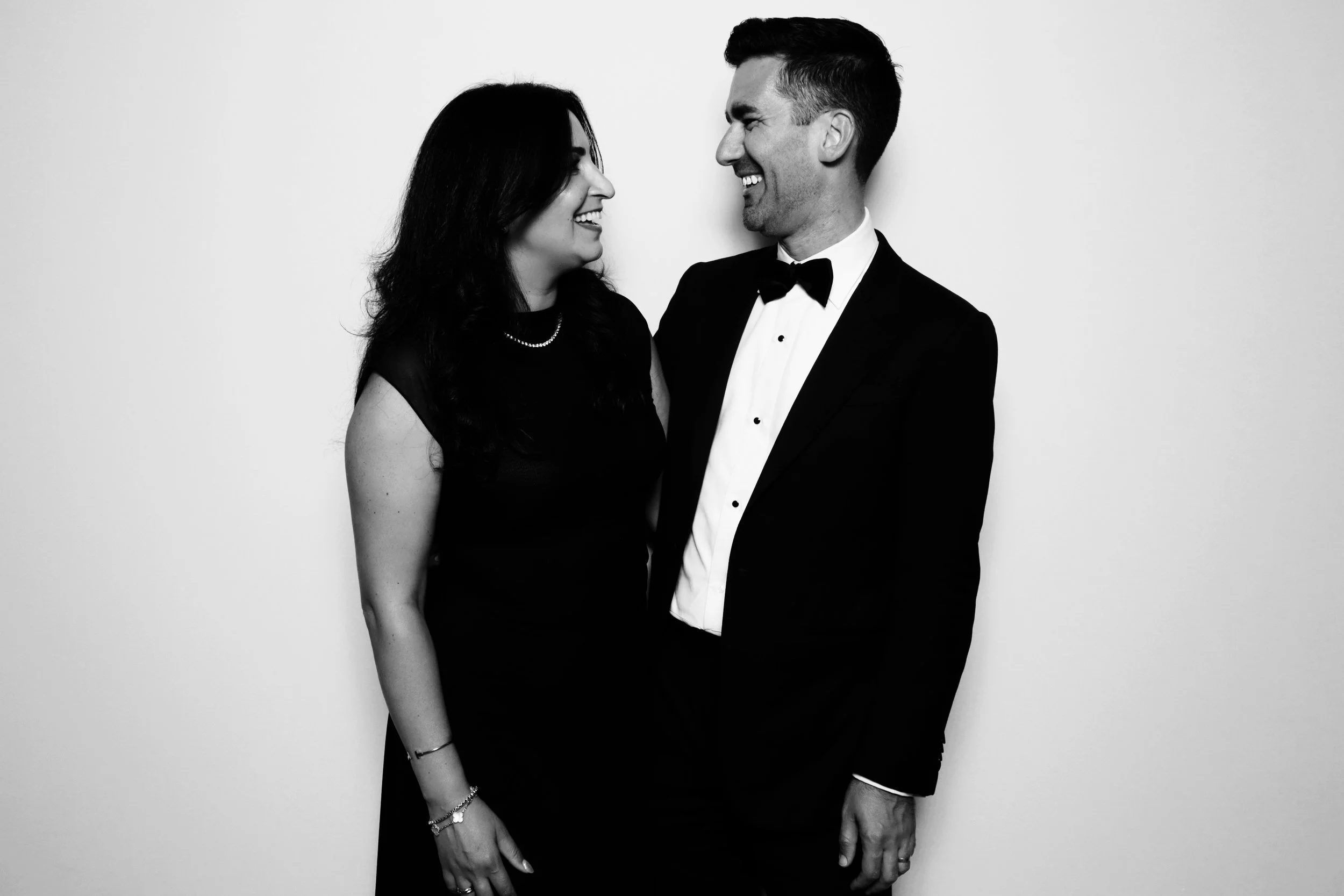 A man and woman dressed in formal attire smiling at each other, standing against a plain background.