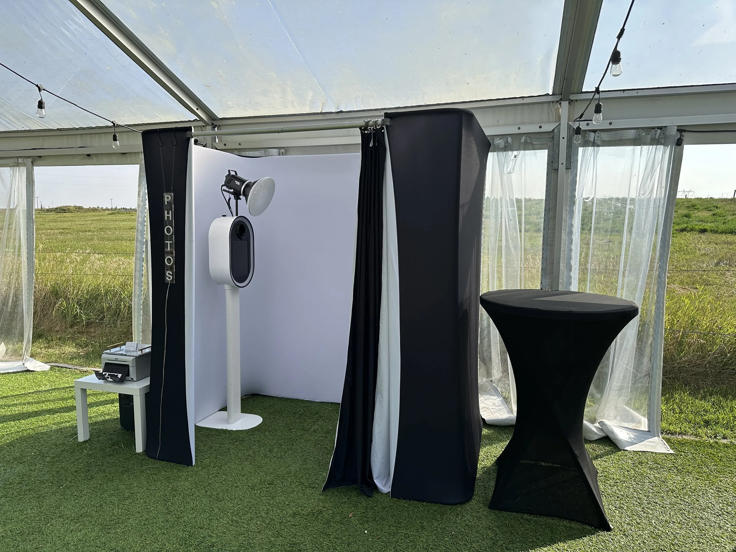 Photo booth setup inside a tent on artificial grass, with a photo booth machine, a light with a softbox, a photo booth background, a small white table with photo strips, a tall black curtain, a black cocktail table, and sheer curtains with a grassy f