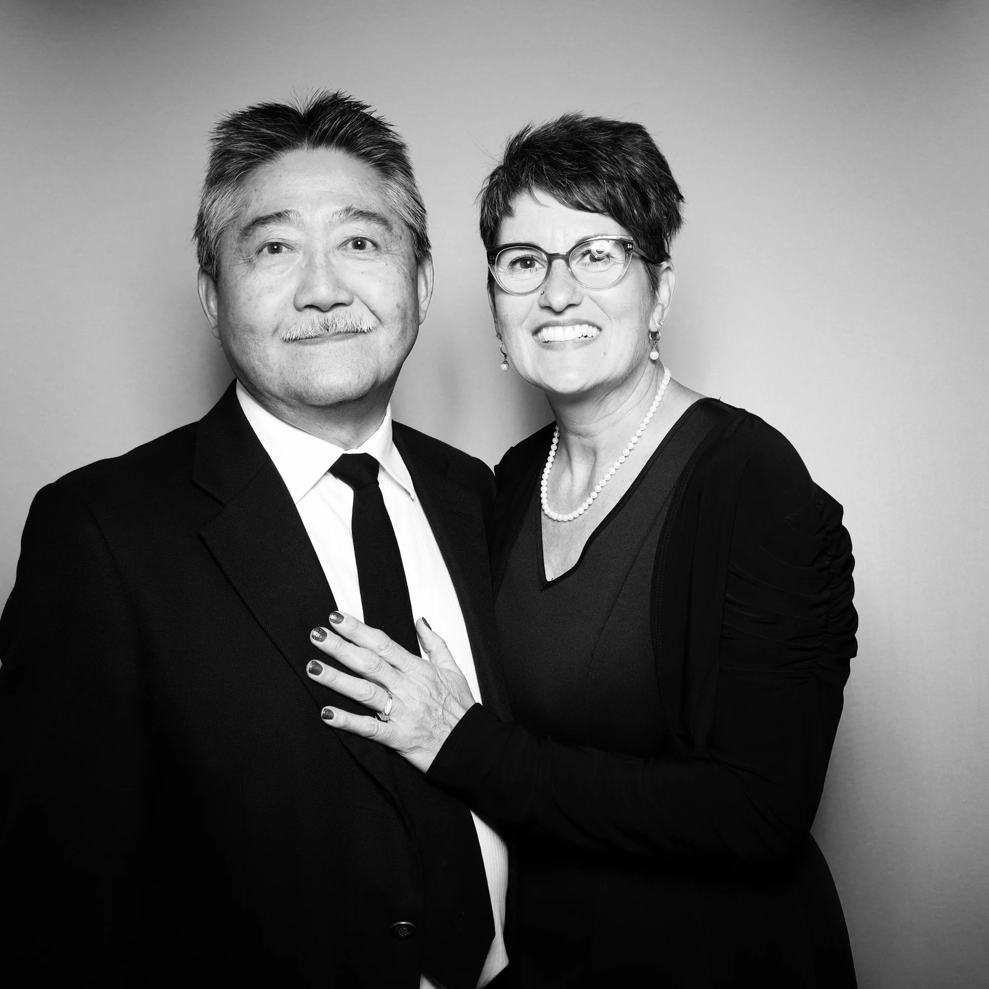 A black and white photo of a man in a suit and a woman in glasses smiling, with the woman touching the man's chest with her hand.