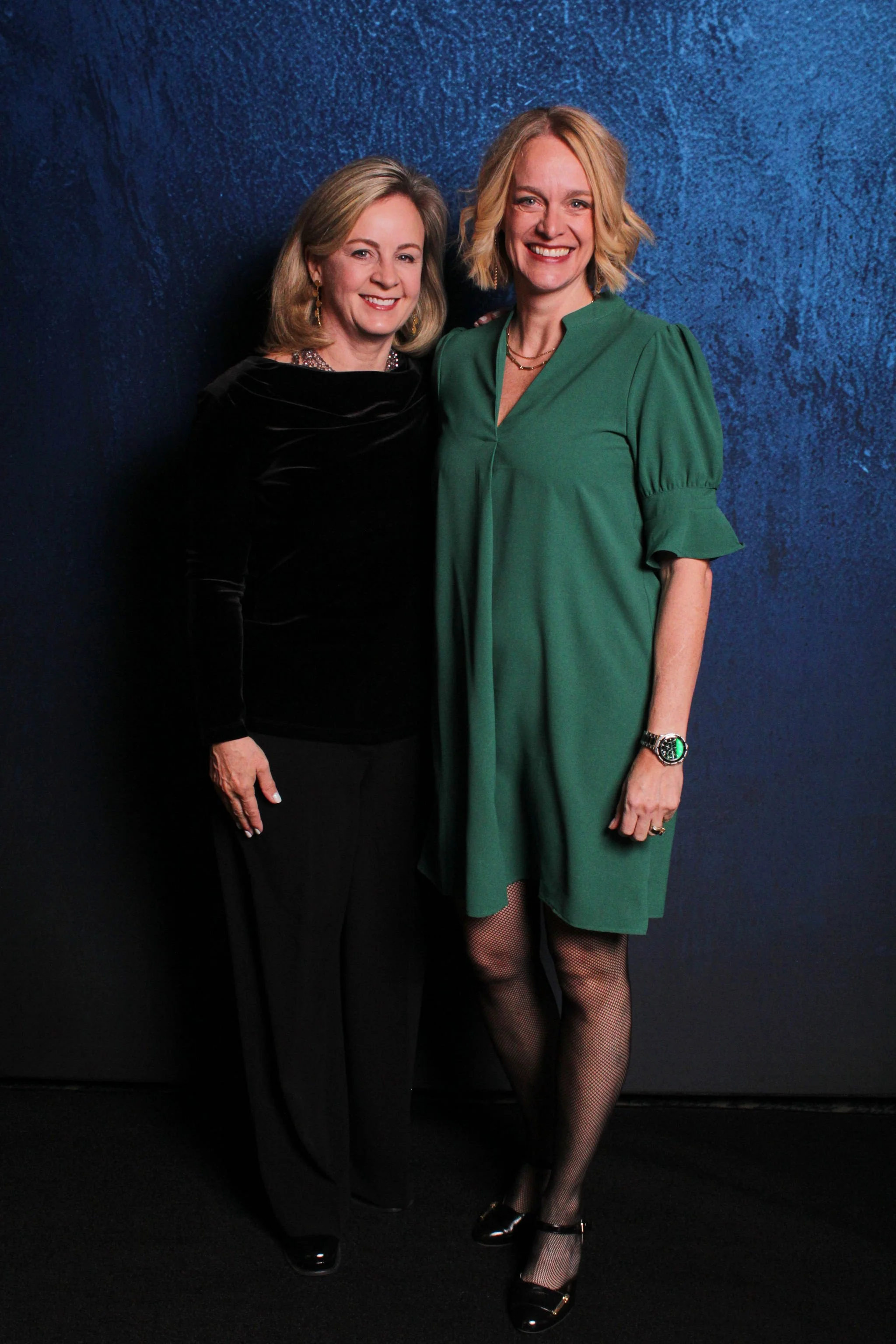 Two women standing together, smiling, in front of a blue textured background. The woman on the left is wearing a black velvet top and black pants, while the woman on the right is wearing a green dress with ruffled sleeves and fishnet stockings.