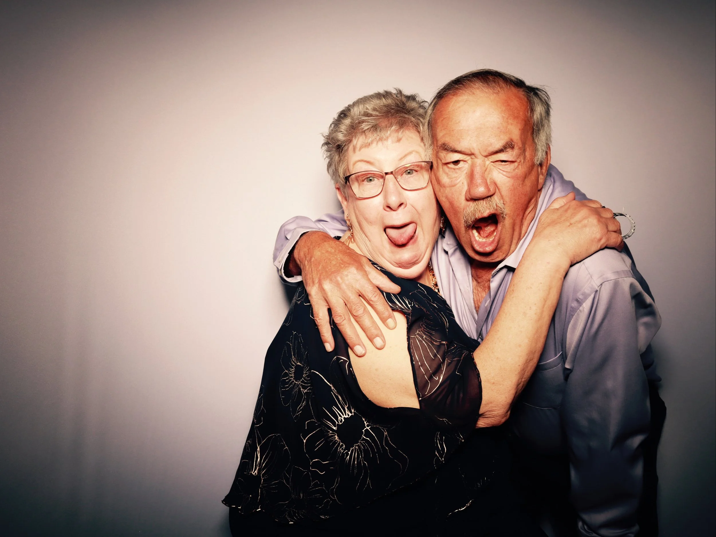 An elderly woman and man hugging each other and making silly faces in front of a plain background.