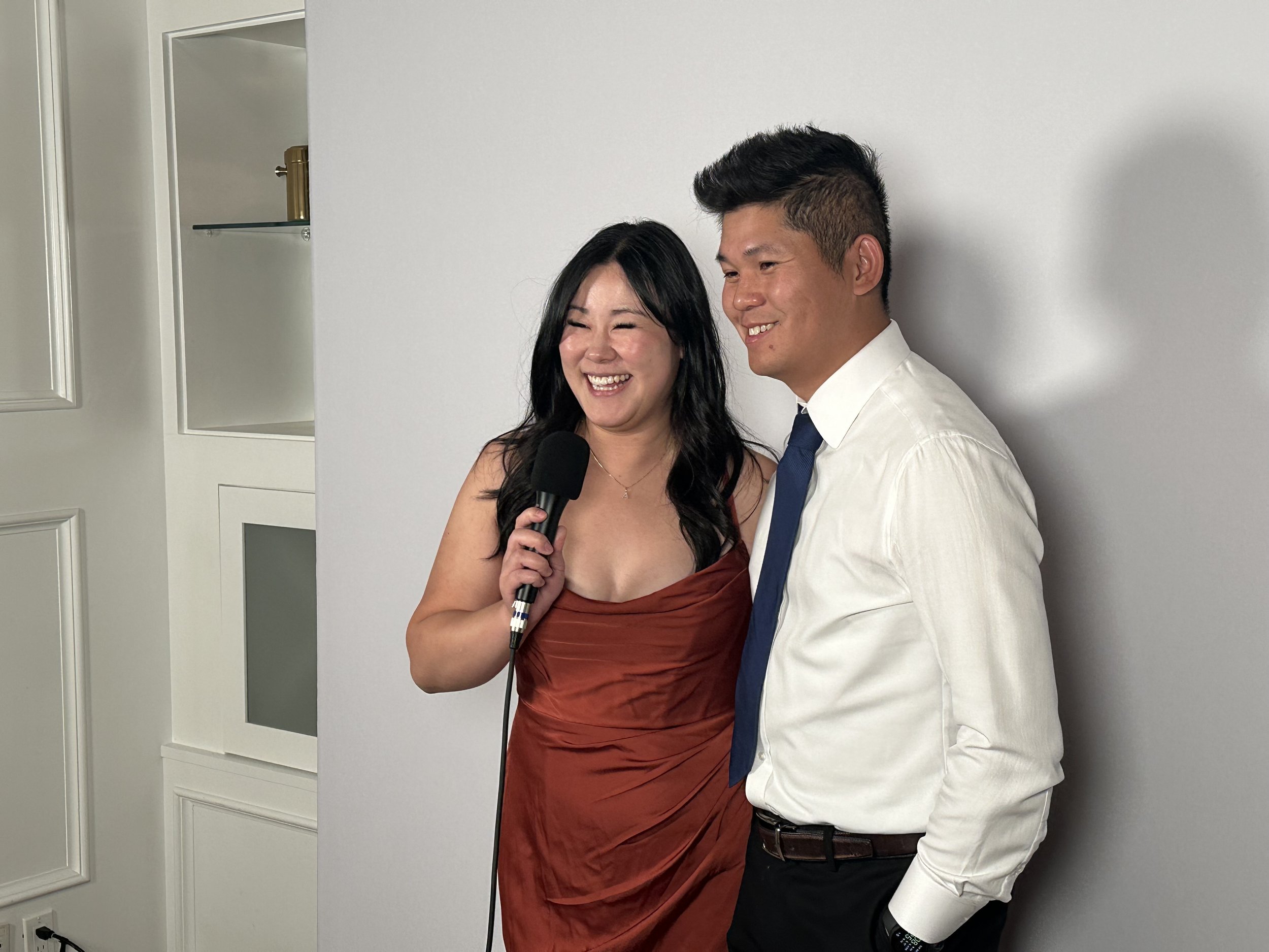 A woman in a rust-colored dress and a man in a white shirt and navy tie stand together, smiling, with the woman holding a microphone, against a plain white wall.