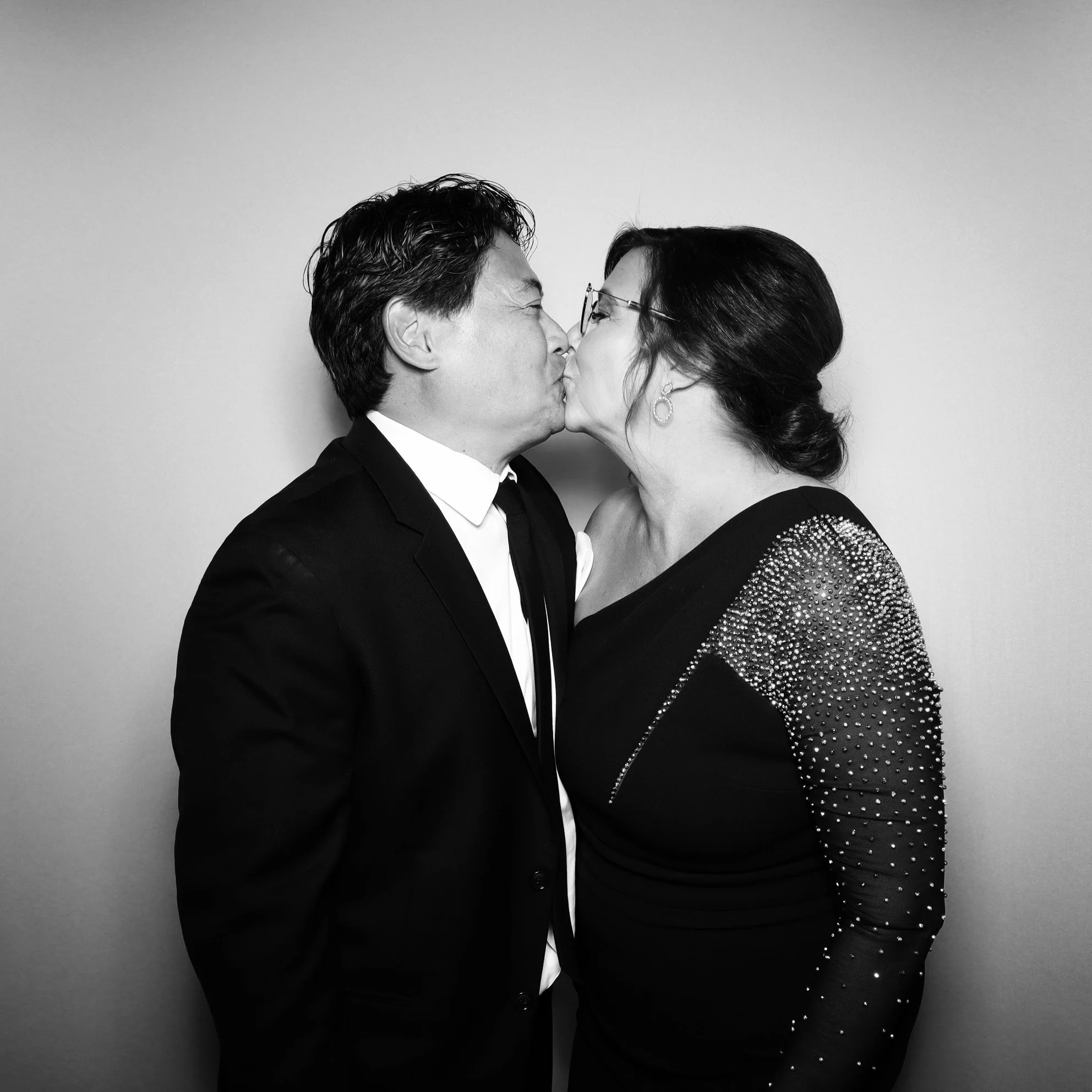 A black and white photo of a man and woman sharing a kiss, with the man dressed in a suit and the woman wearing a dress with rhinestone embellishments on the sleeves.