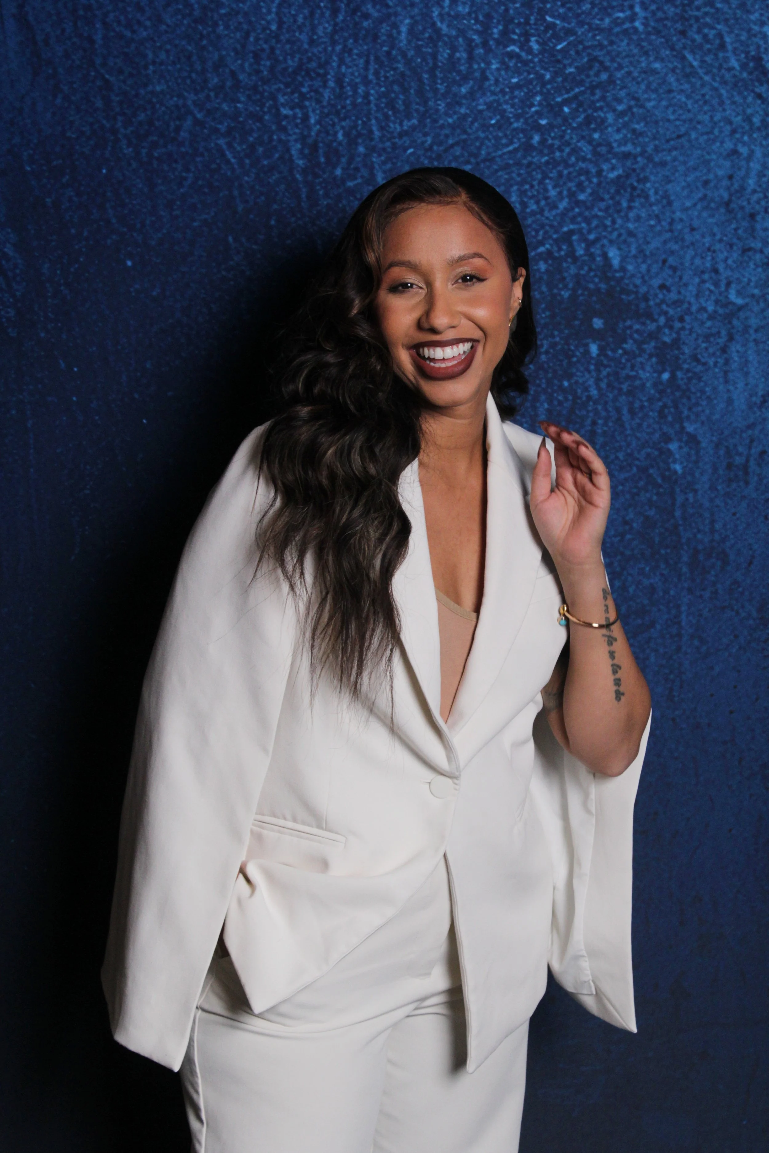 A woman with long wavy black hair, wearing a white suit, smiling confidently against a blue textured background.