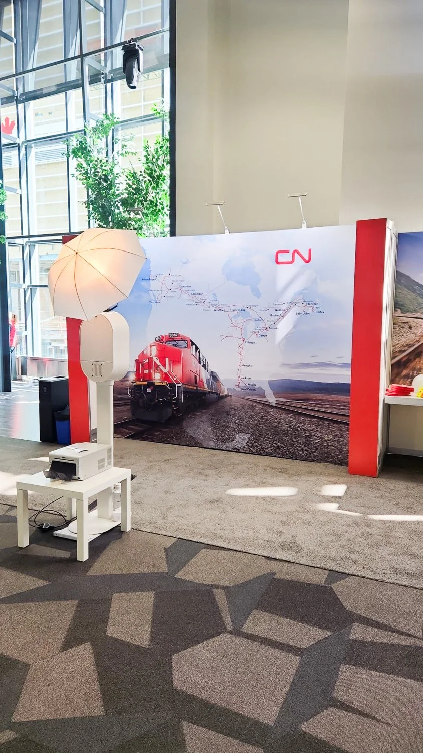Indoor exhibit featuring CN railroad artwork with a red train on a track, a map of train routes, and a large CN logo. Display includes a sunshade umbrella and a projector on a small table, set against a backdrop of glass windows and indoor greenery.