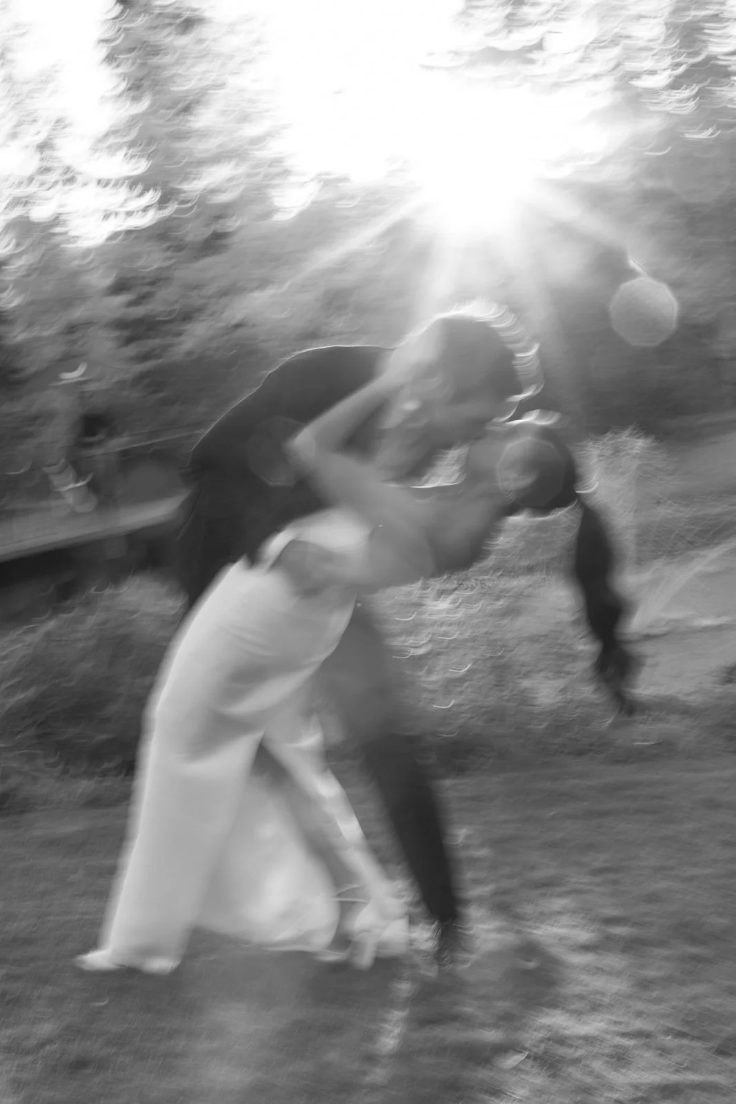 A blurry black and white photo of a couple kissing near a body of water, with the sun shining brightly overhead.