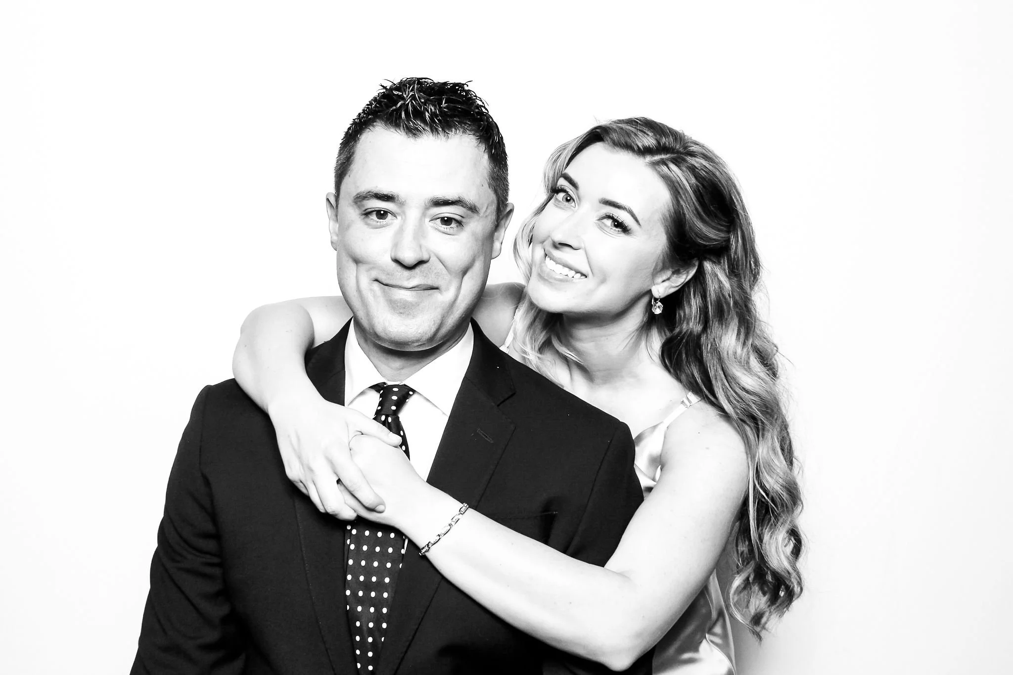 A black-and-white photo of a smiling man in a suit and woman in a dress embracing and looking at the camera.