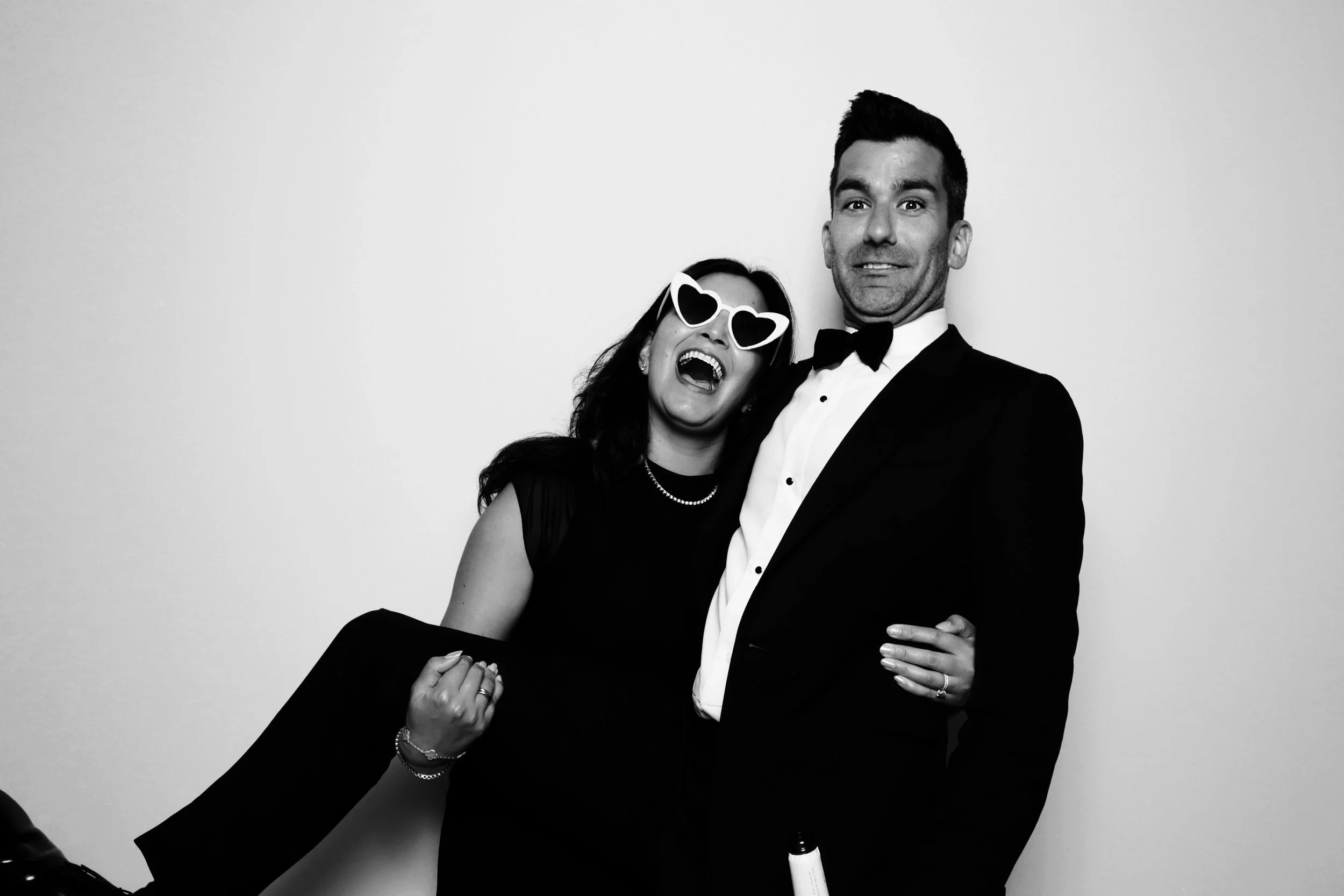 A man in a tuxedo holding a woman in a black dress and heart-shaped sunglasses, both smiling and celebrating at a formal event.