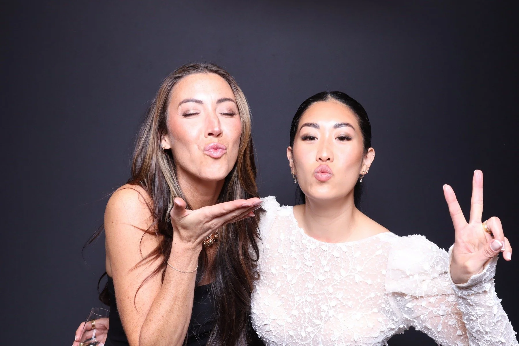 Two women in a photo against a dark background, blowing kisses and making peace signs.