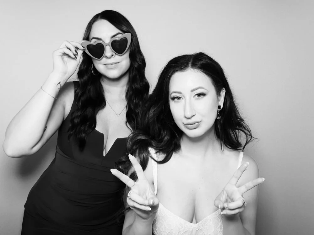 Two women posing in a photo booth, one wearing heart-shaped sunglasses and the other making a peace sign.