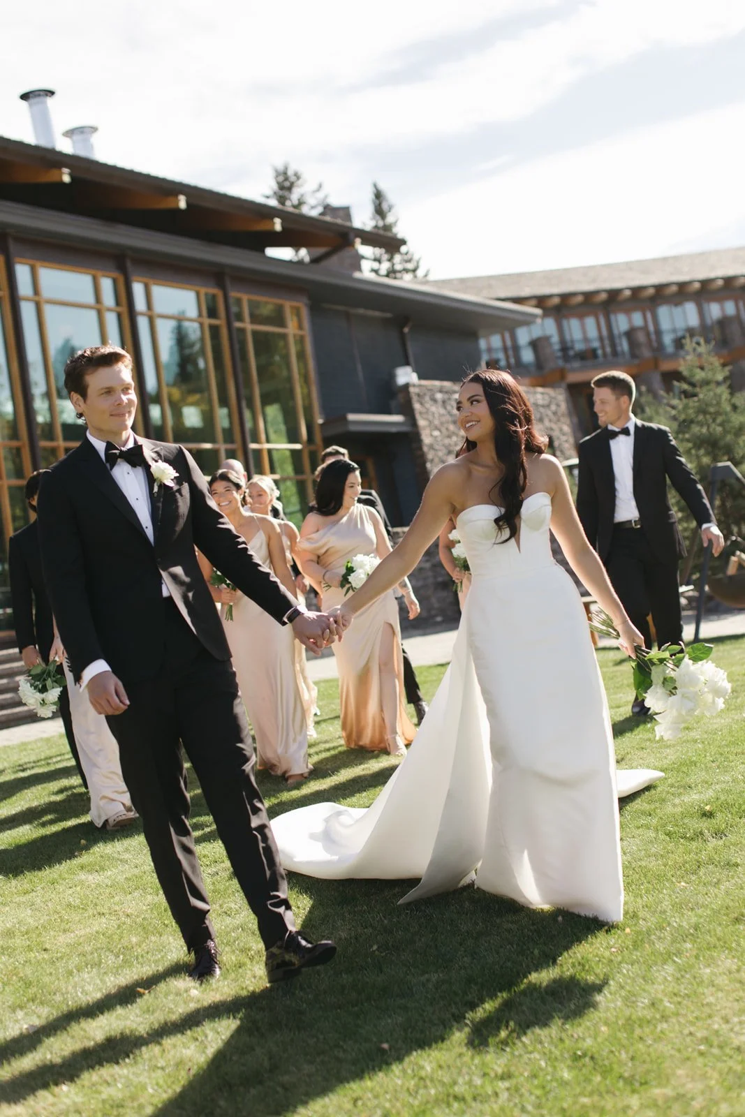A wedding party outdoors, including a bride and groom holding hands, with bridesmaids and groomsmen in the background, on a grassy area in front of a modern house with large windows.