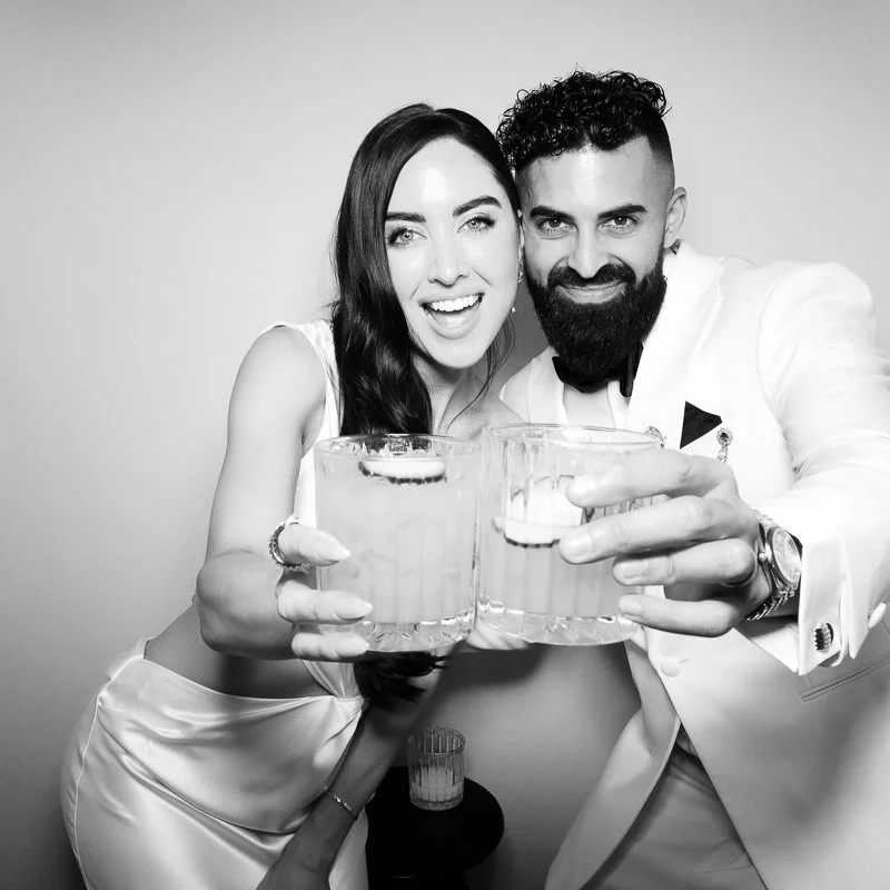 A smiling woman and man dressed in formal attire, holding glasses of drinks for a toast, against a plain background.