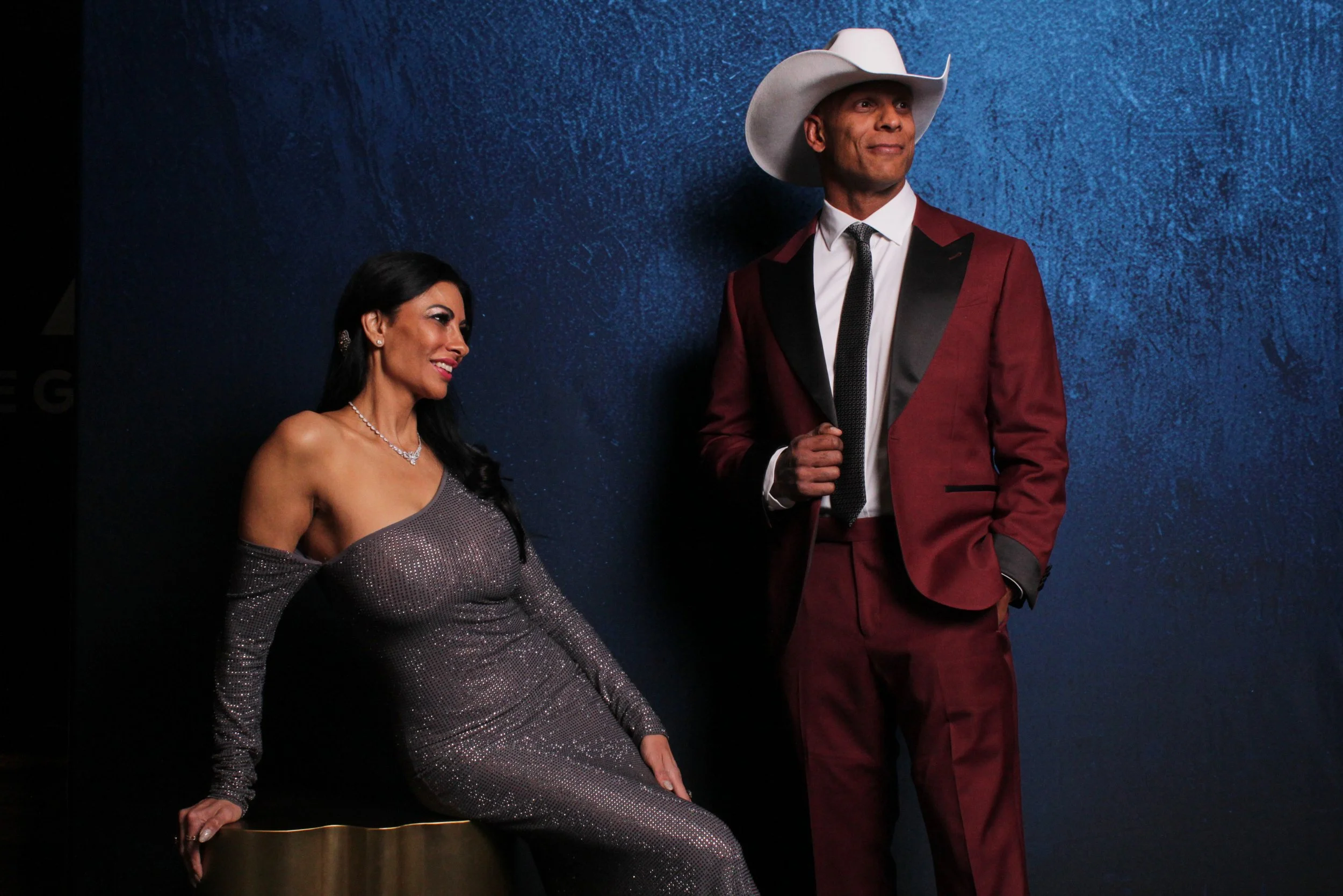 A woman in a glittery, one-shoulder dress sitting on a gold-colored surface next to a man in a red tuxedo with black lapels, white shirt, black tie, and a white cowboy hat. They are in front of a dark blue textured background.