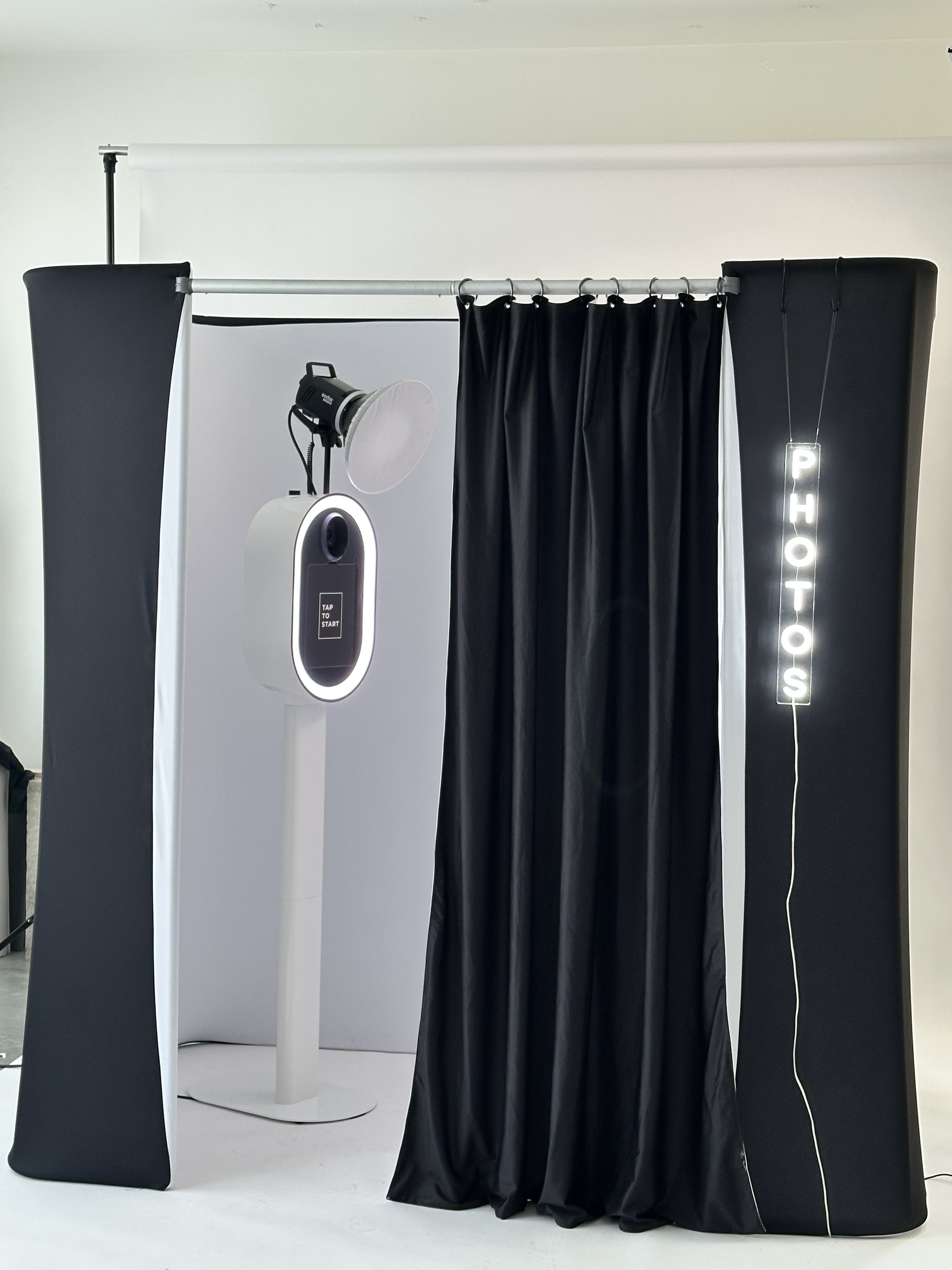Photo booth with black curtains, a camera, and a neon 'PHOTOS' sign.