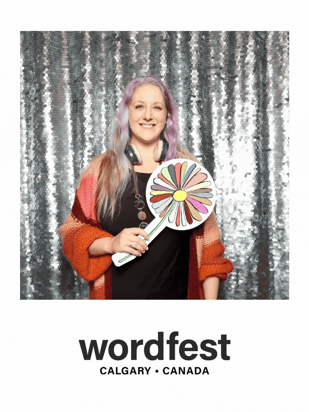 A woman with light purple and blonde hair, wearing a colorful orange and red sweater, smiling, holding a colorful flower cutout, standing in front of a silver sequin backdrop, with the word 'wordfest' and 'Calgary Canada' printed below.