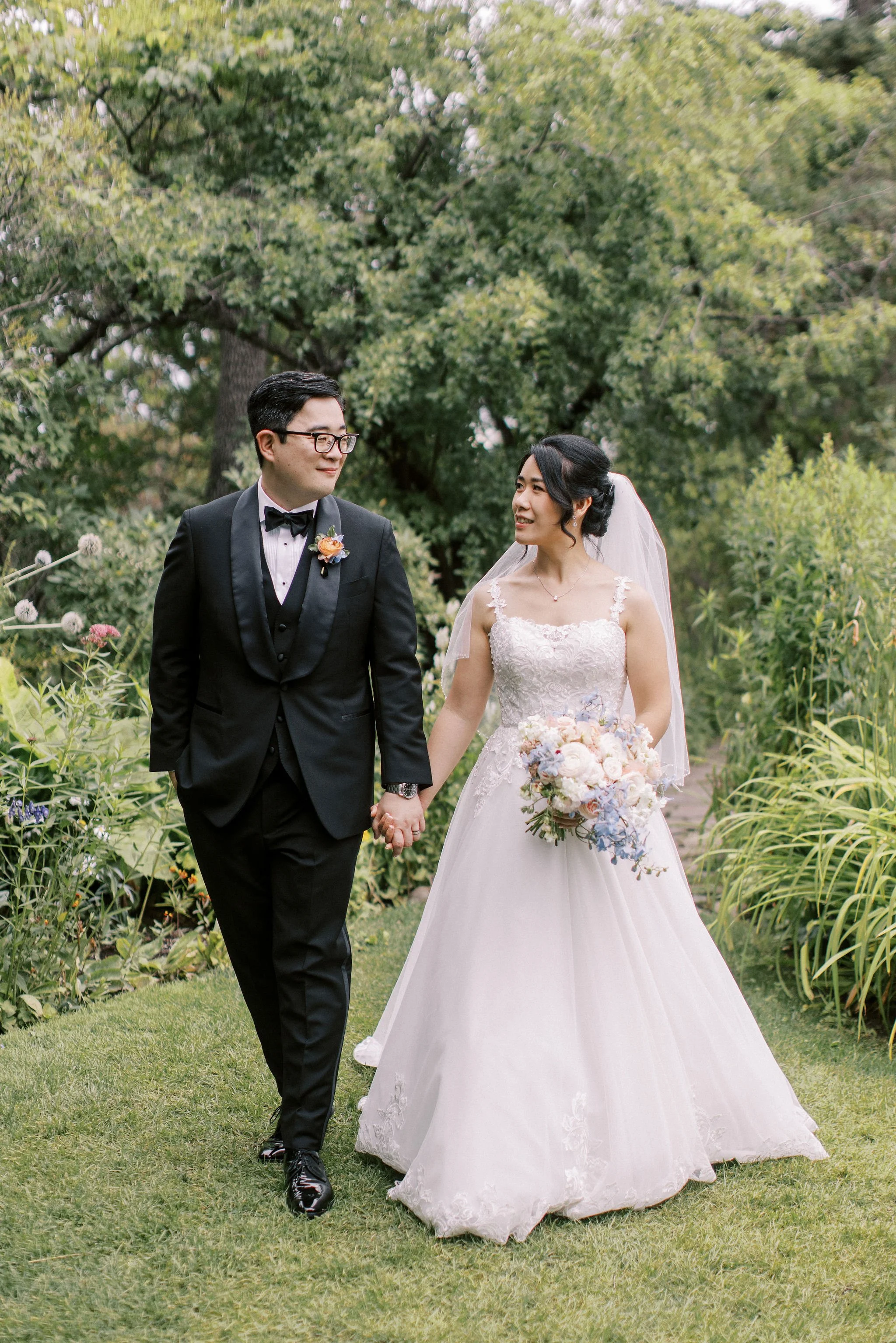 A bride and groom holding hands and smiling at each other in a garden. The groom wears a black tuxedo and bowtie, glasses, and a boutonniere. The bride wears a white lace wedding gown and holds a bouquet of flowers. They are surrounded by greenery an