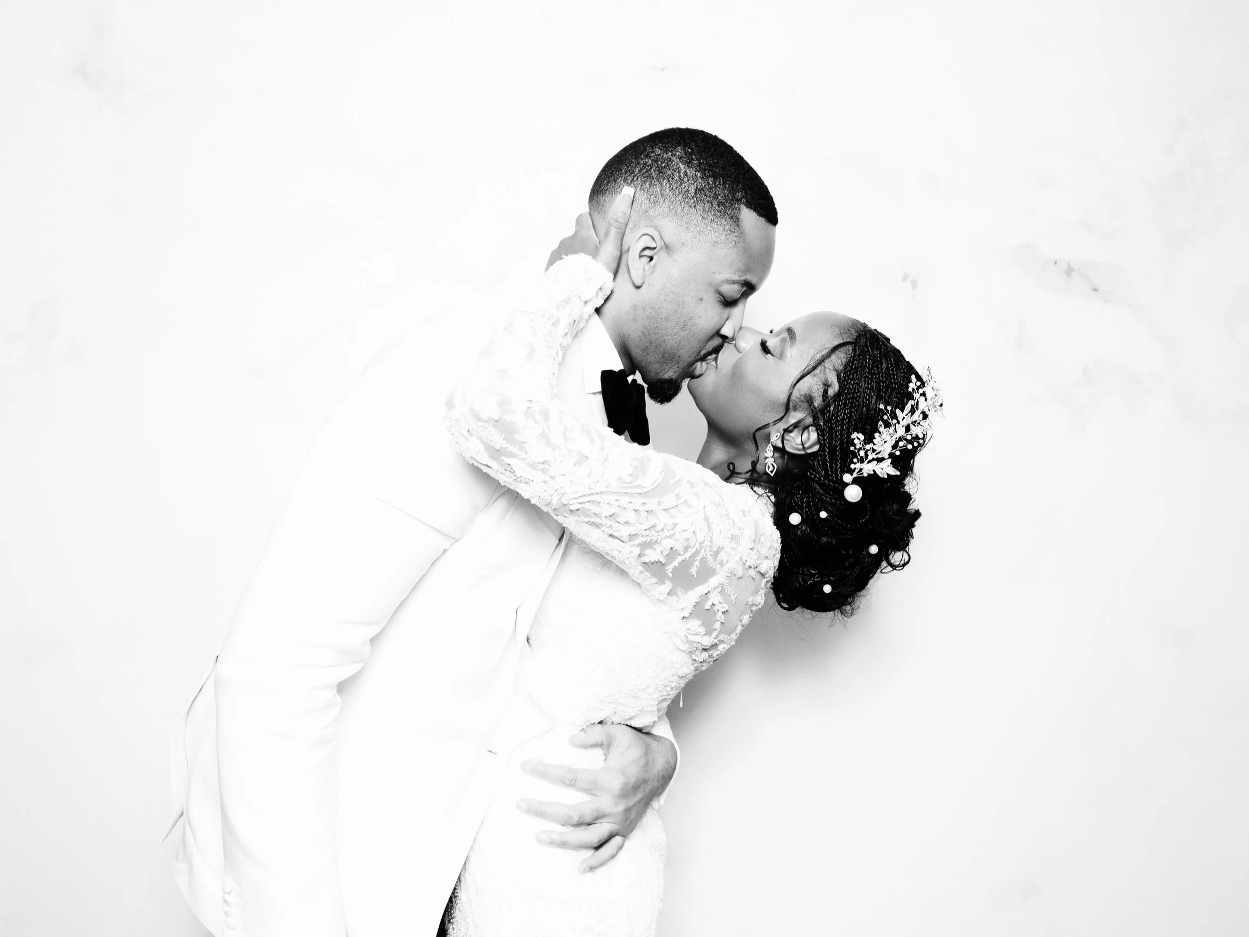 A black and white photo of a bride and groom sharing a kiss, with the groom wearing a white tuxedo and the bride in a lace gown with intricate hair jewelry.