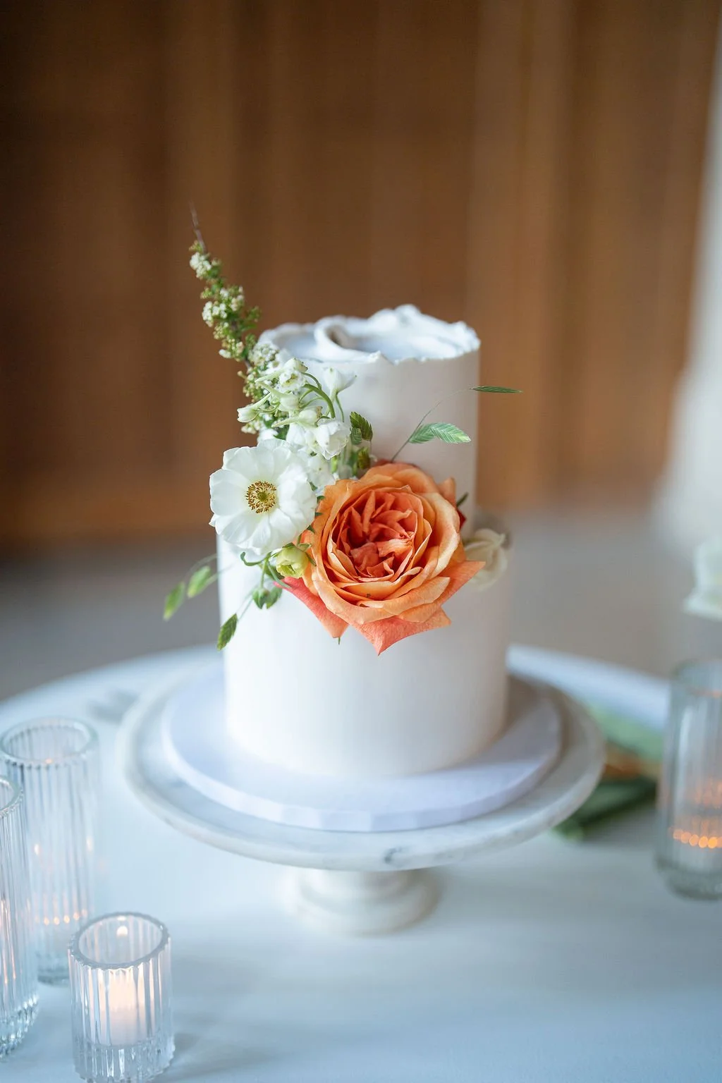 A white, round wedding cake decorated with fresh flowers including a peach-colored rose and white blossoms, placed on a white marble cake stand.