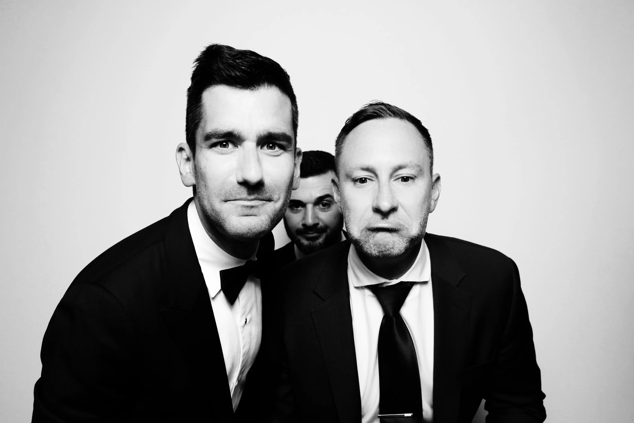 Three men in suits, two with bow ties and one with a tie, posing in front of a plain background, black and white photo.