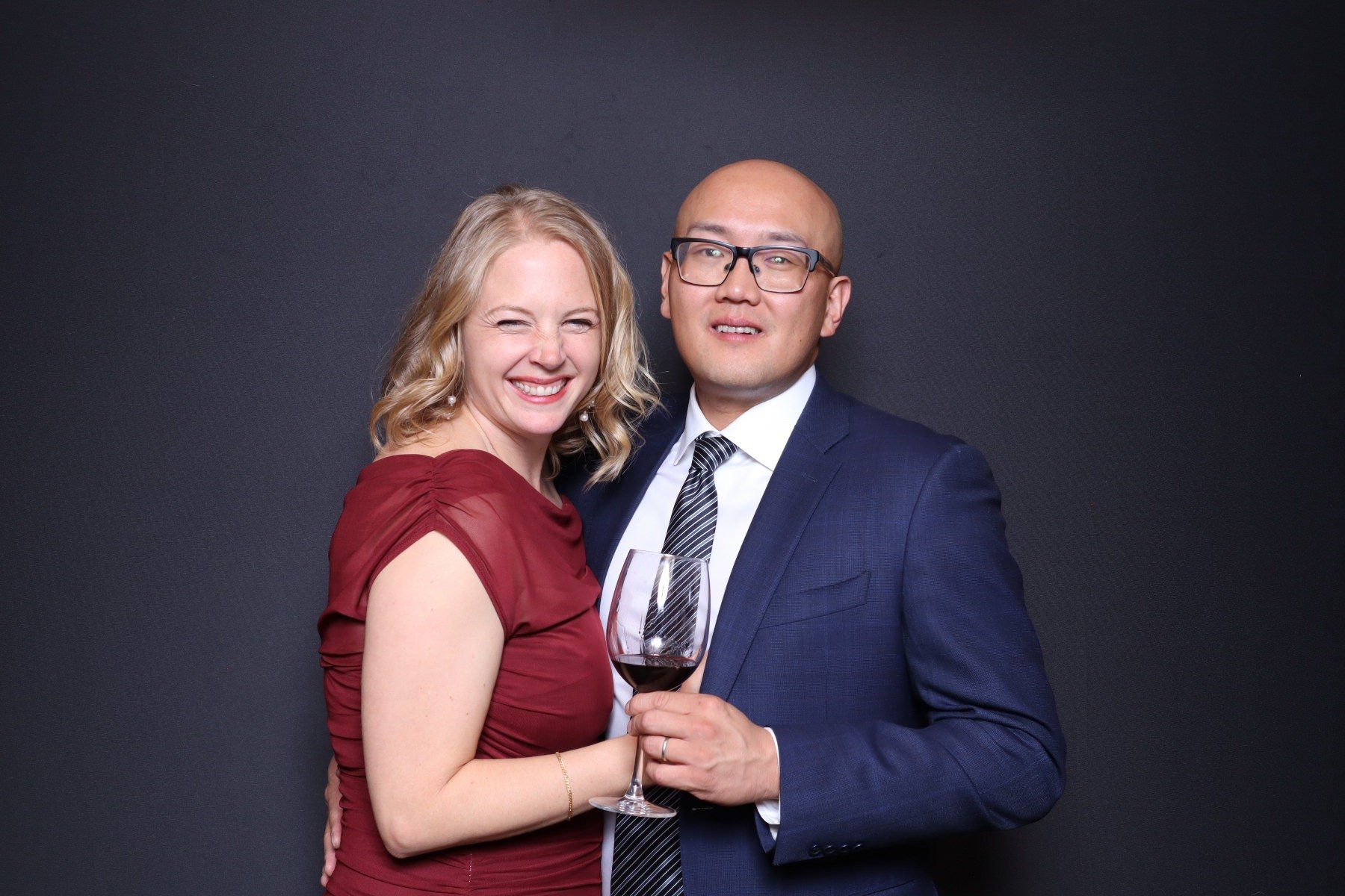 A smiling woman with blonde hair in a red dress holding a glass of red wine, standing next to a man in a blue suit and tie holding a glass of red wine, against a dark background.