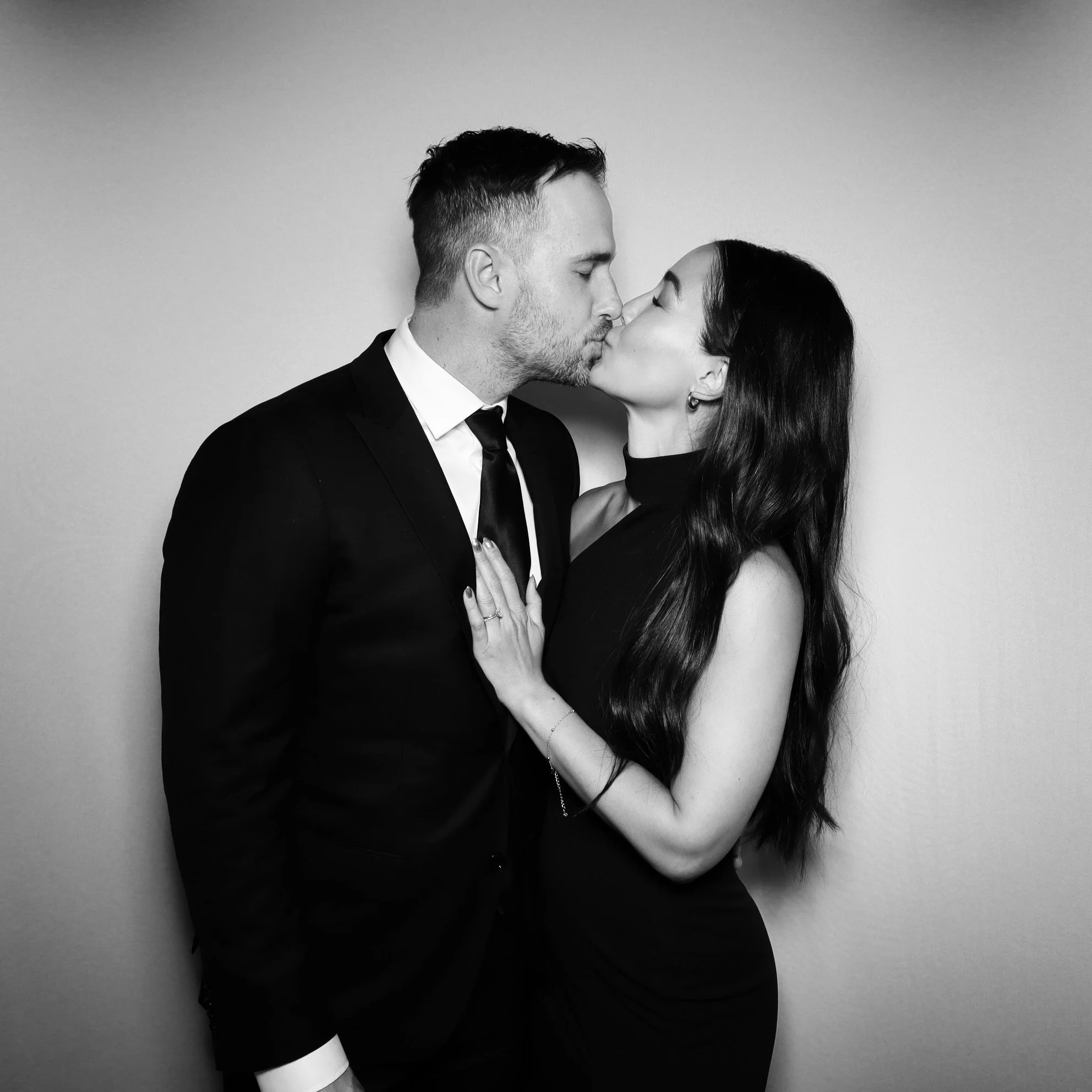 A black-and-white photo of a man and woman kissing, dressed elegantly, against a plain light-colored background.