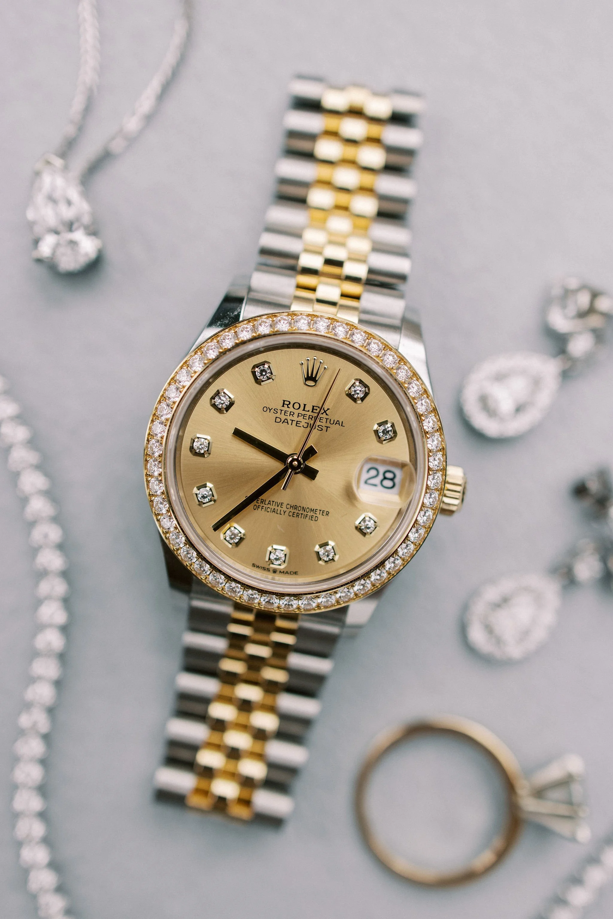 A gold and silver Rolex Oyster Perpetual Datejust wristwatch with diamond accents on the bezel and dial, displaying the date 28, surrounded by jewelry including bracelets, necklaces, and rings.