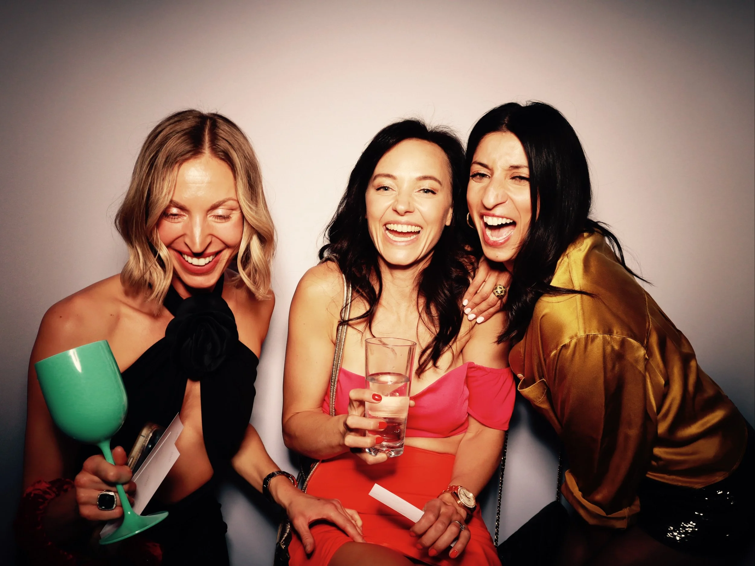 Three women laughing and enjoying drinks at a party or celebration, against a plain background.