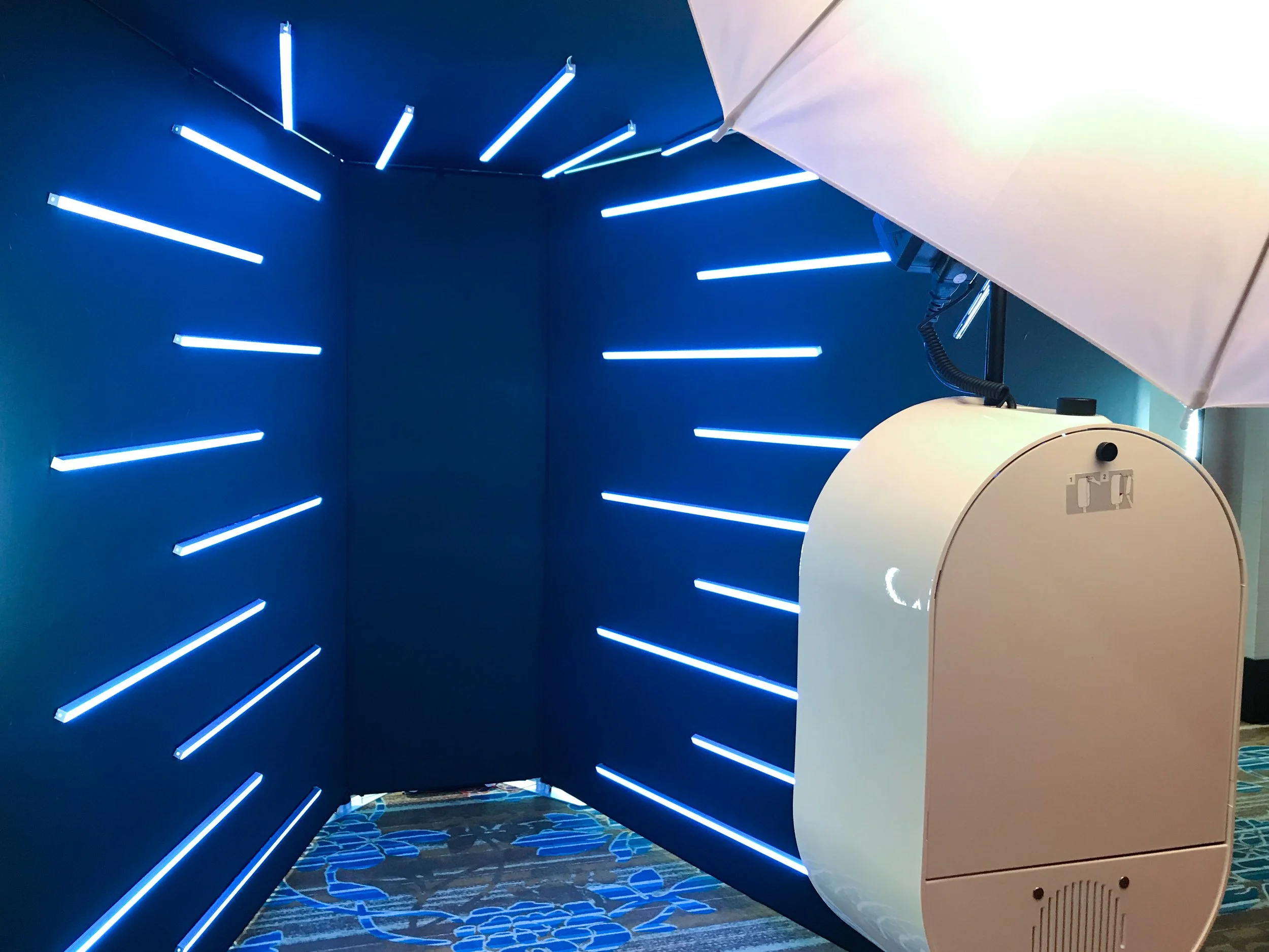 A photo of a dark blue photo booth with horizontal LED light strips and a white umbrella with a inside light, attached to the booth. The floor has a patterned carpet.