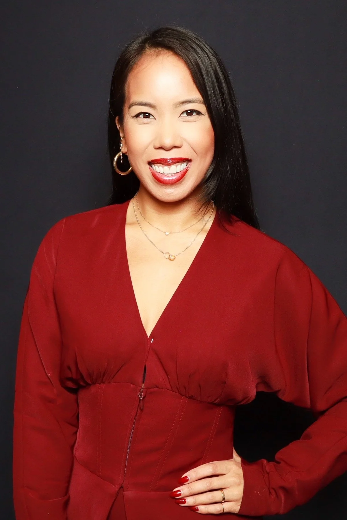 A woman with long black hair, wearing a red dress, smiling and posing against a dark background.
