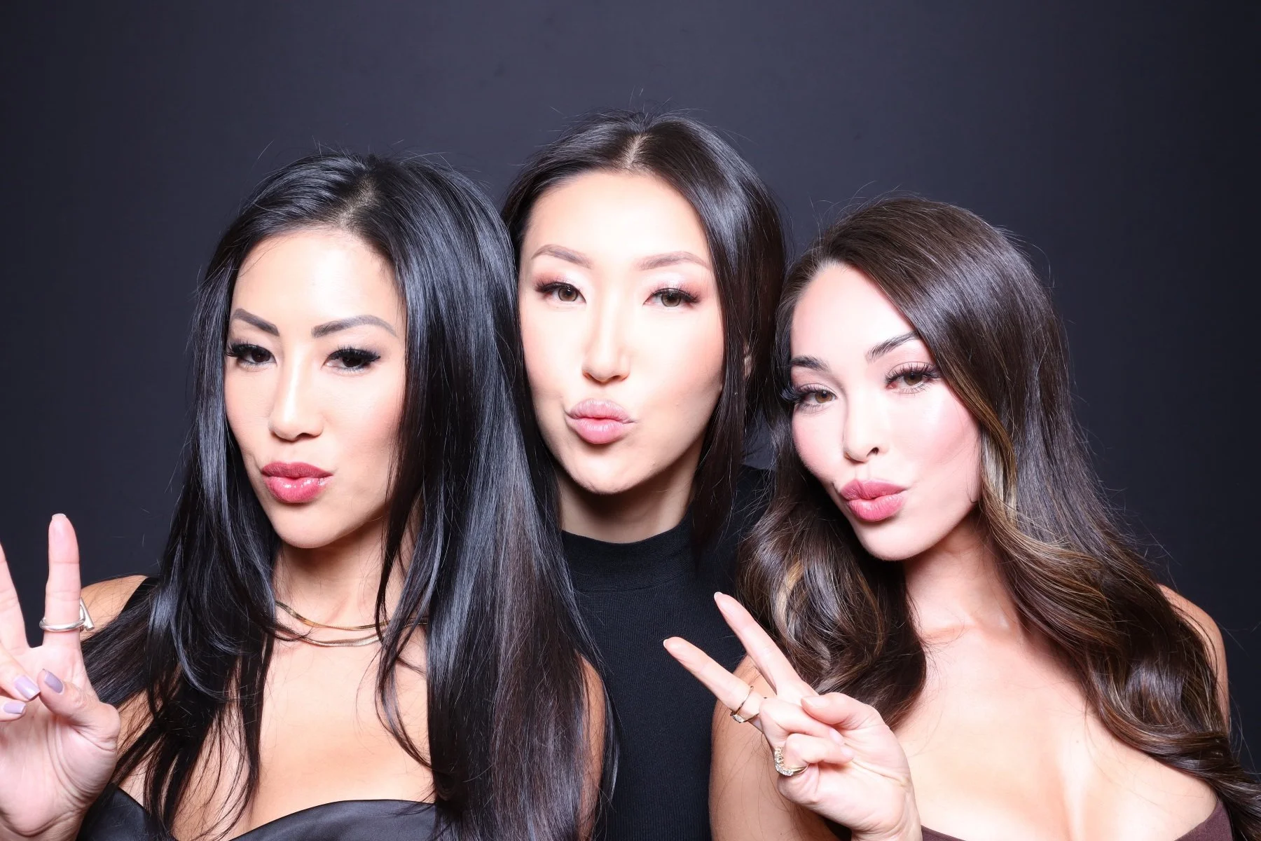 Three women with long, dark hair posing together against a dark background, making peace signs and puckering their lips.