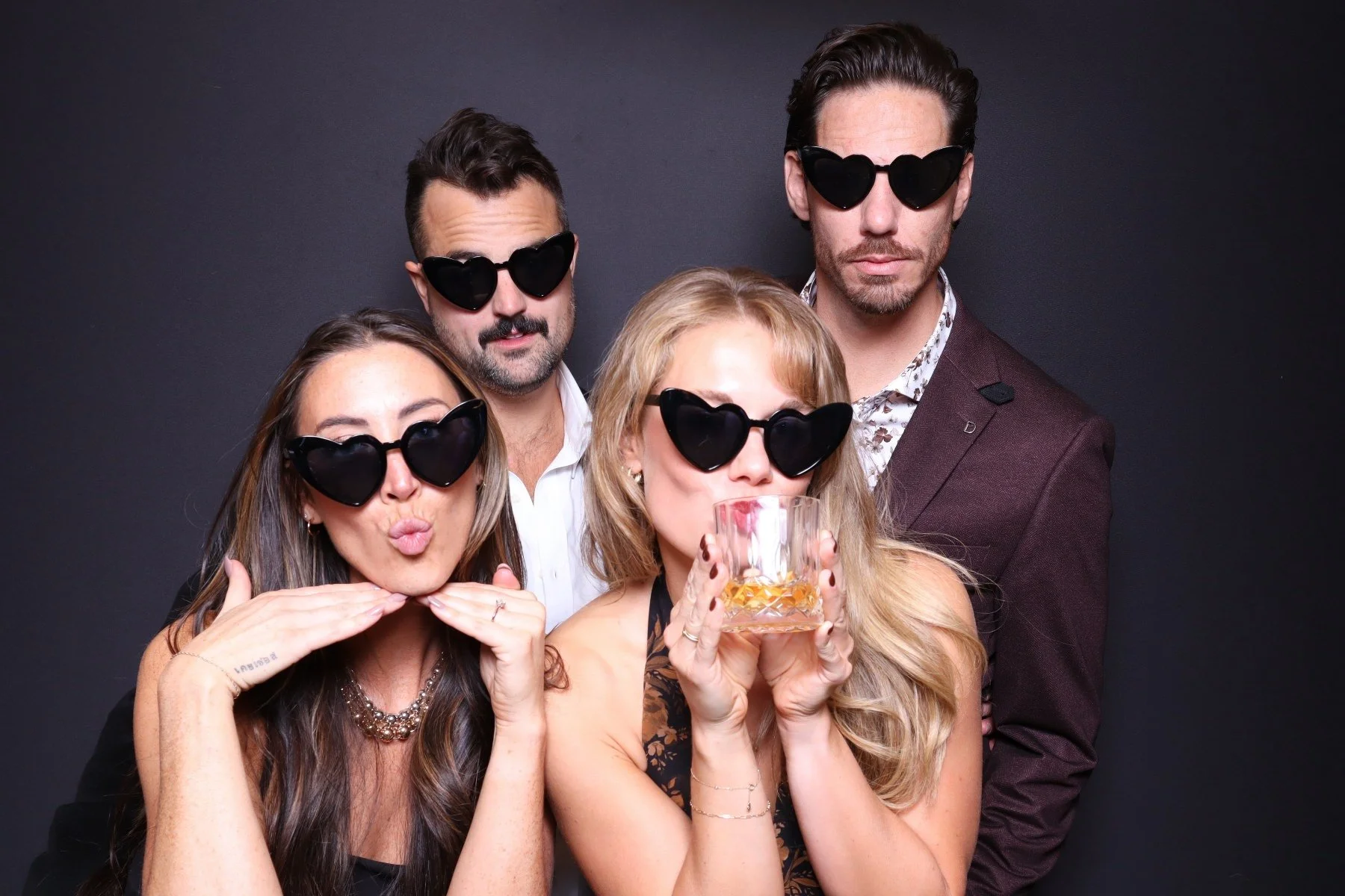 Group of five people wearing black heart-shaped sunglasses, posing against a dark background, with one woman holding a glass of whiskey.
