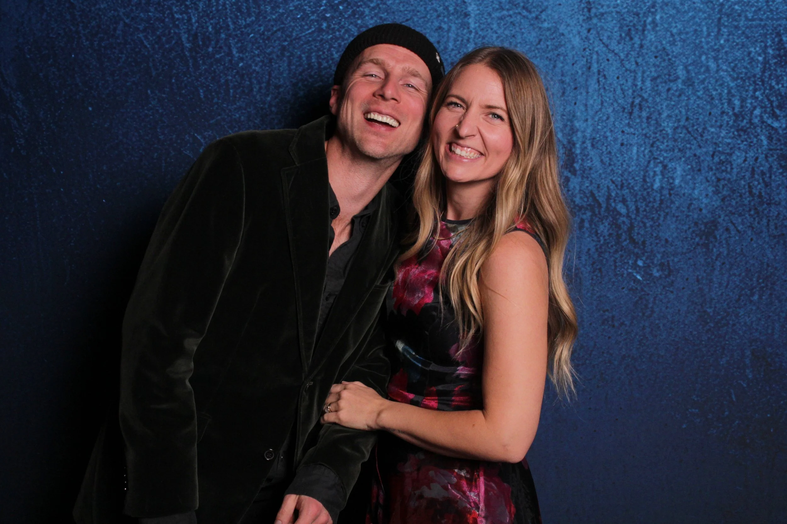 A man and woman standing together, smiling and posing for a photo, with a dark blue textured background.