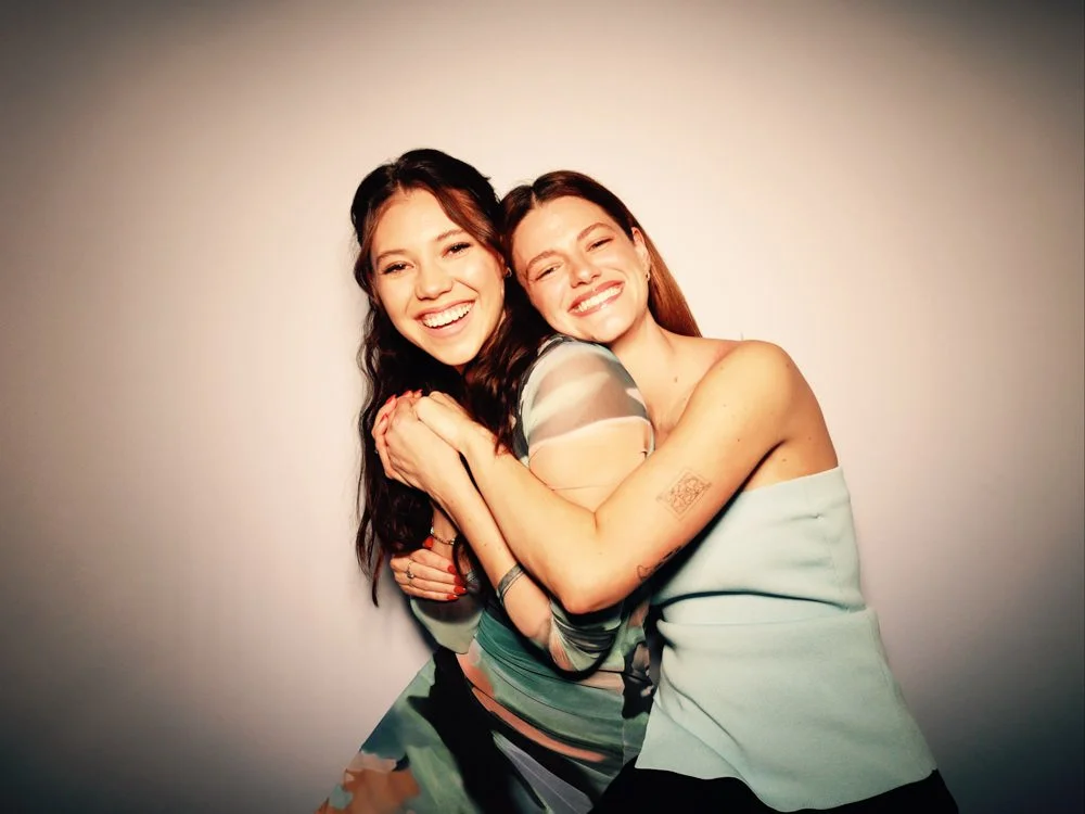 Two women smiling and hugging each other against a plain background.
