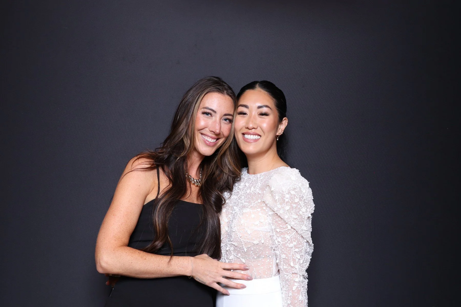 Two women smiling and standing close together against a plain dark background.