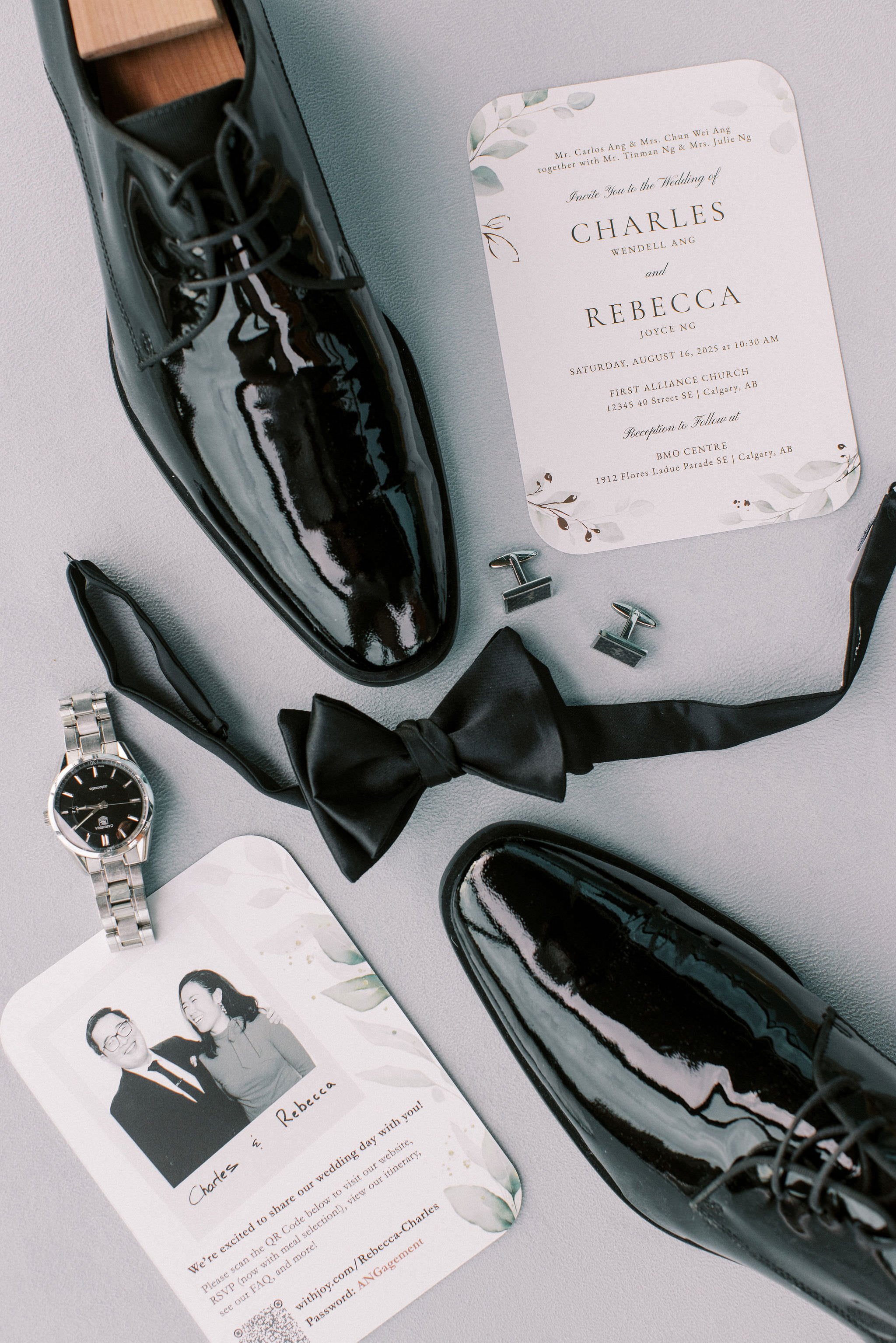 Wedding invitation, black shiny dress shoes, a black bow tie, a silver wristwatch, a wedding RSVP card with a photo and a wedding invitation with text, and two small metal clips on a gray surface.