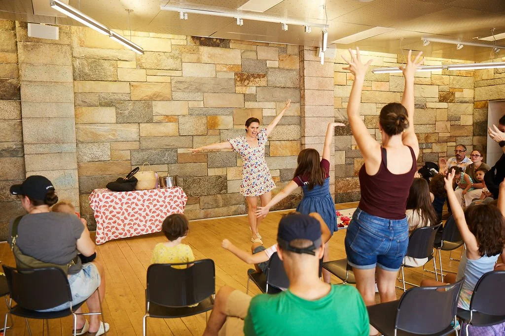A woman with brown hair stands in front of an audience with her arms wide and standing on one leg. Members of the audience are standing up to copy her pose.