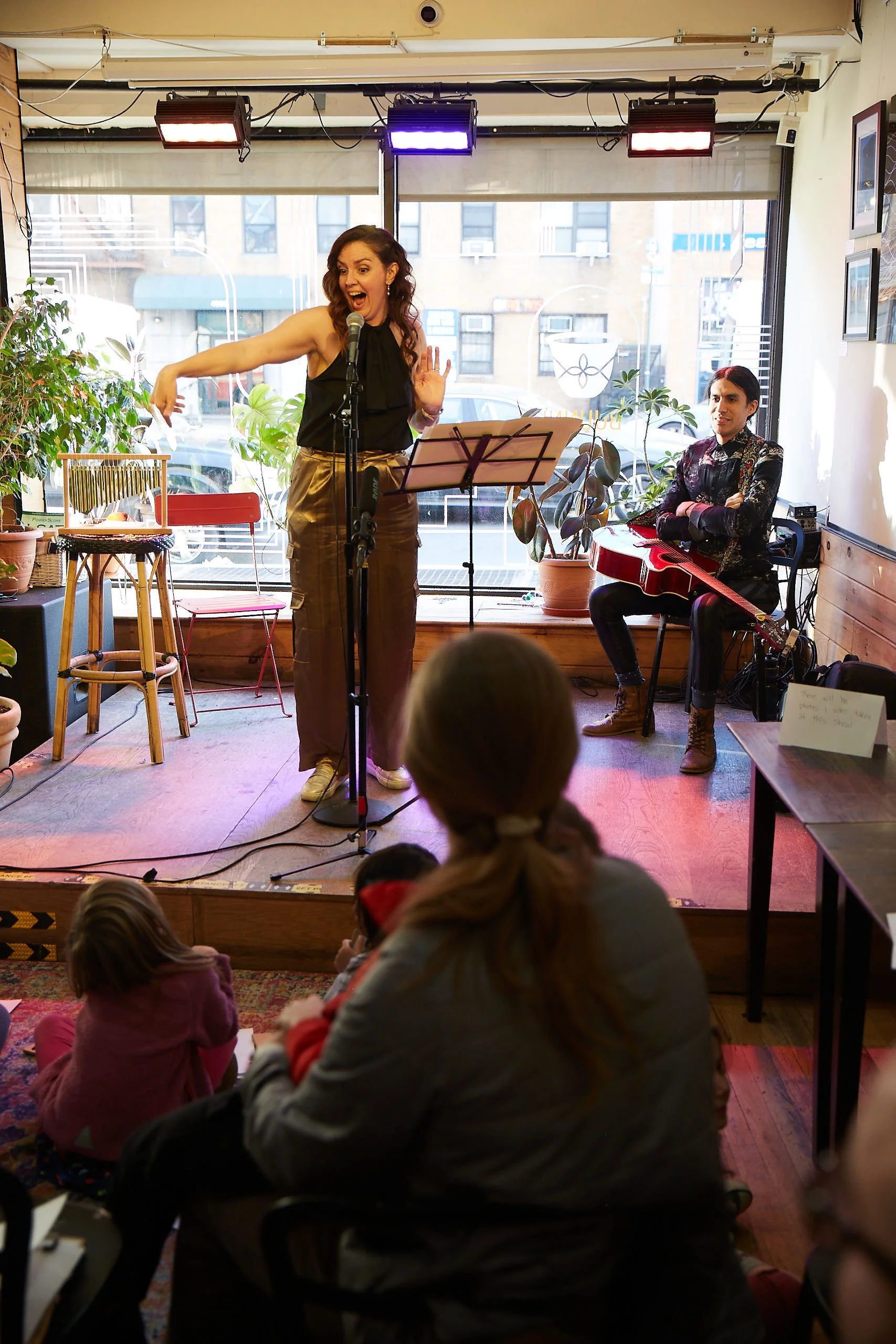 A woman talking passionately into a microphone on a small stage in a cafe, with a musician sitting beside her playing a red guitar. The audience, including a woman with red hair and children, watches the live performance.