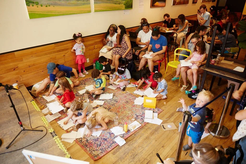 Children and adults gathered in an art or reading studio, with children sitting on the rug and adults on chairs, engaging in reading and activities, surrounded by books and art on the walls.