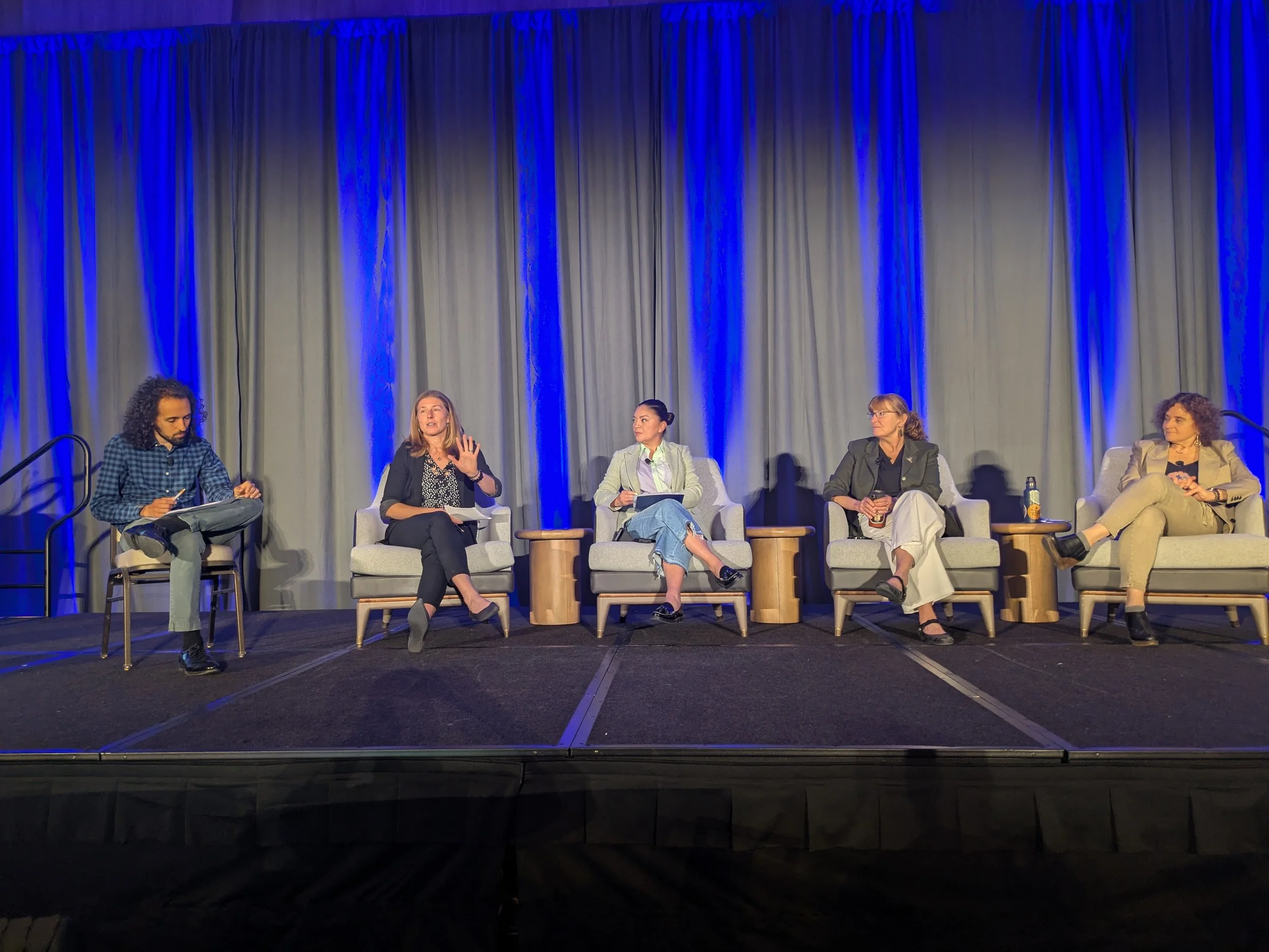 A panel of five speakers seated on stage in front of gray curtains, participating in a plenary discussion. The people are sitting in armchairs with small tables beside them. The stage is lit with blue lighting.