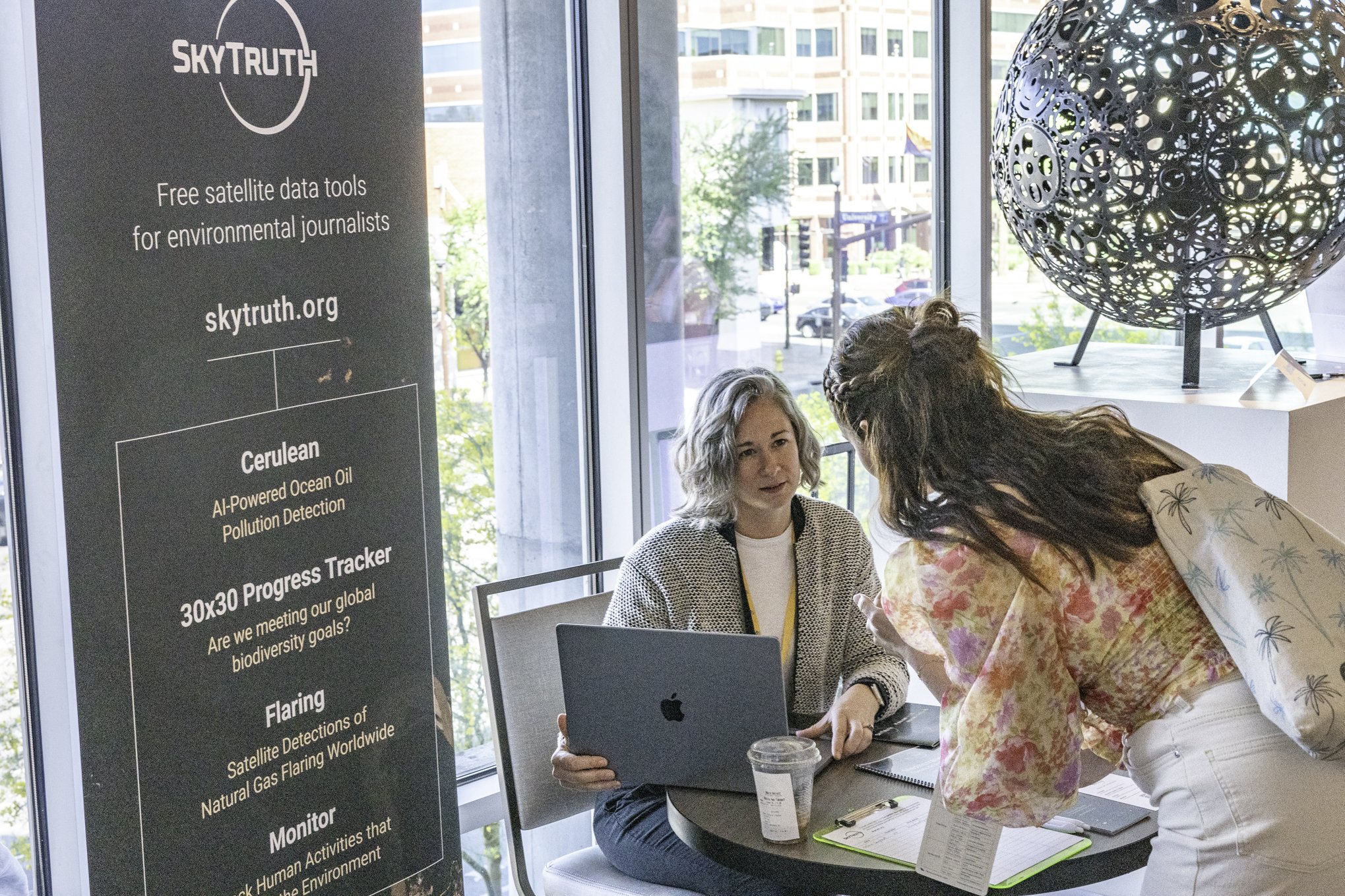 Two women are engaged in a conversation at a table near large windows, with a tall vertical banner advertising Skytruth's environmental data tools in the background. The banner lists services like ocean oil pollution detection and biodiversity goal tracking.