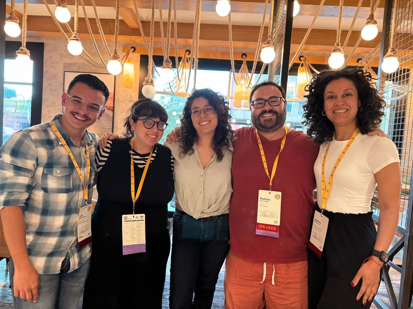 Six people standing together, smiling, in an SEJ2025 conference venue with hanging lights and wooden decor, wearing conference badges.