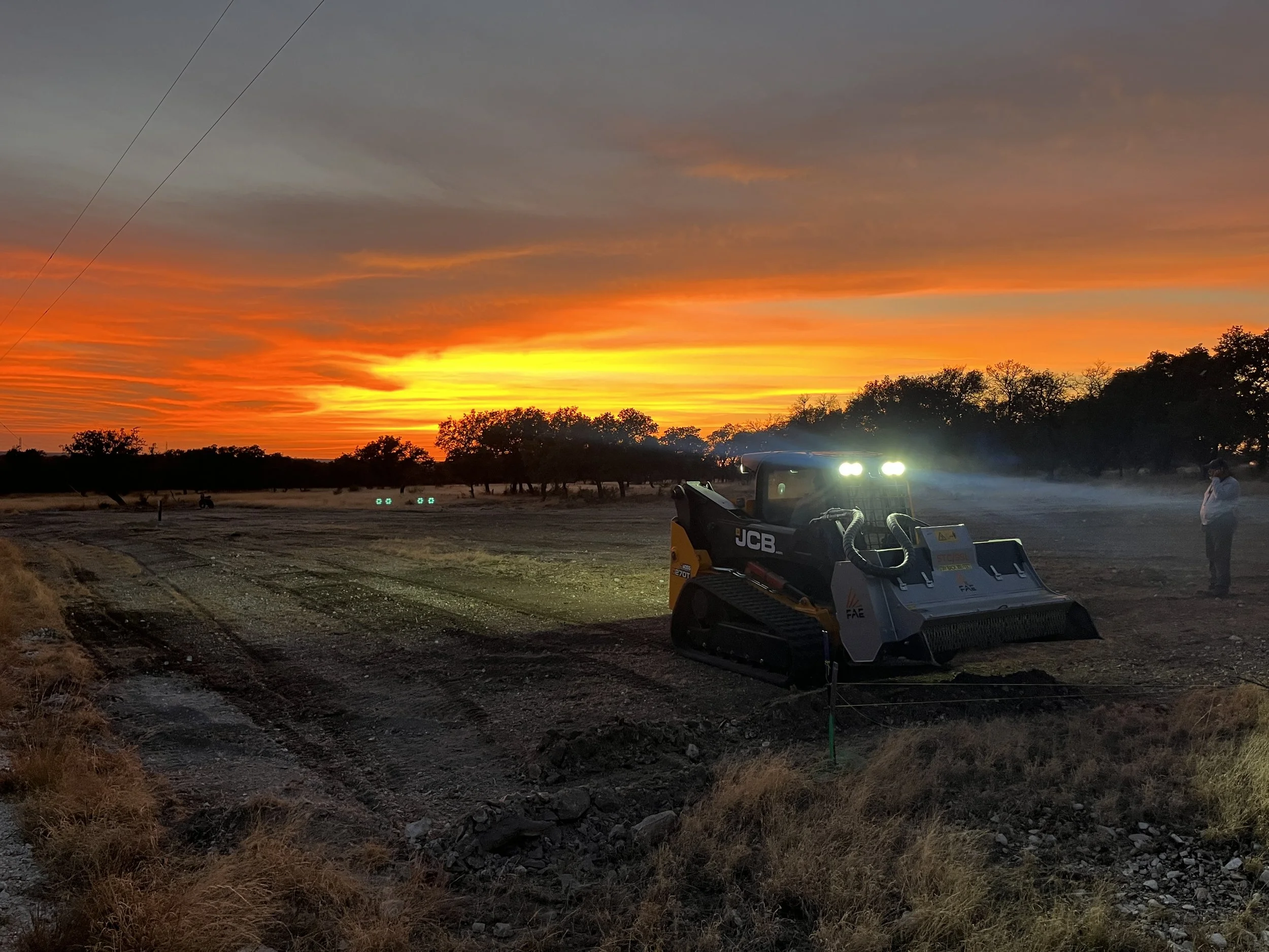 A construction vehicle working at sunset on a dirt road, with a person standing nearby and trees in the background.