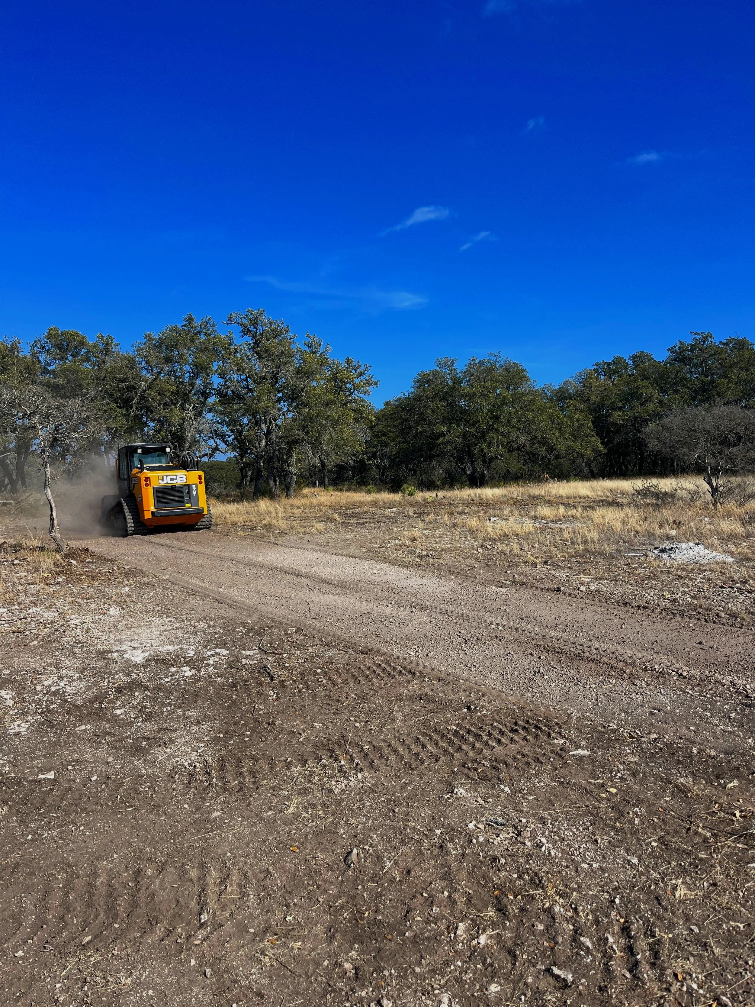 A yellow JCB construction vehicle driving on a dirt road in a rural area with trees and a clear blue sky.