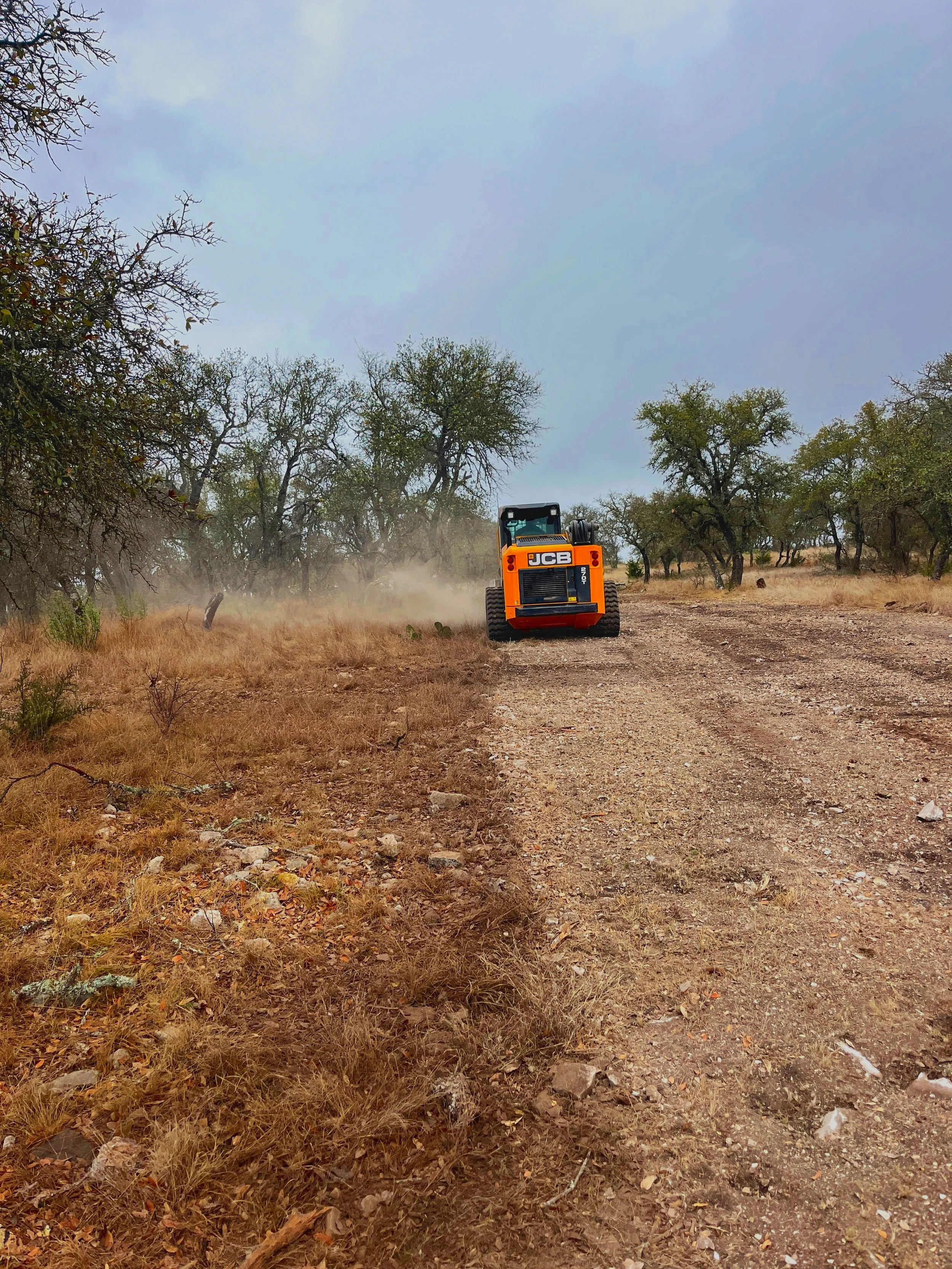 A yellow JCB construction vehicle driving down a dirt road in a rural area with trees and overcast sky.