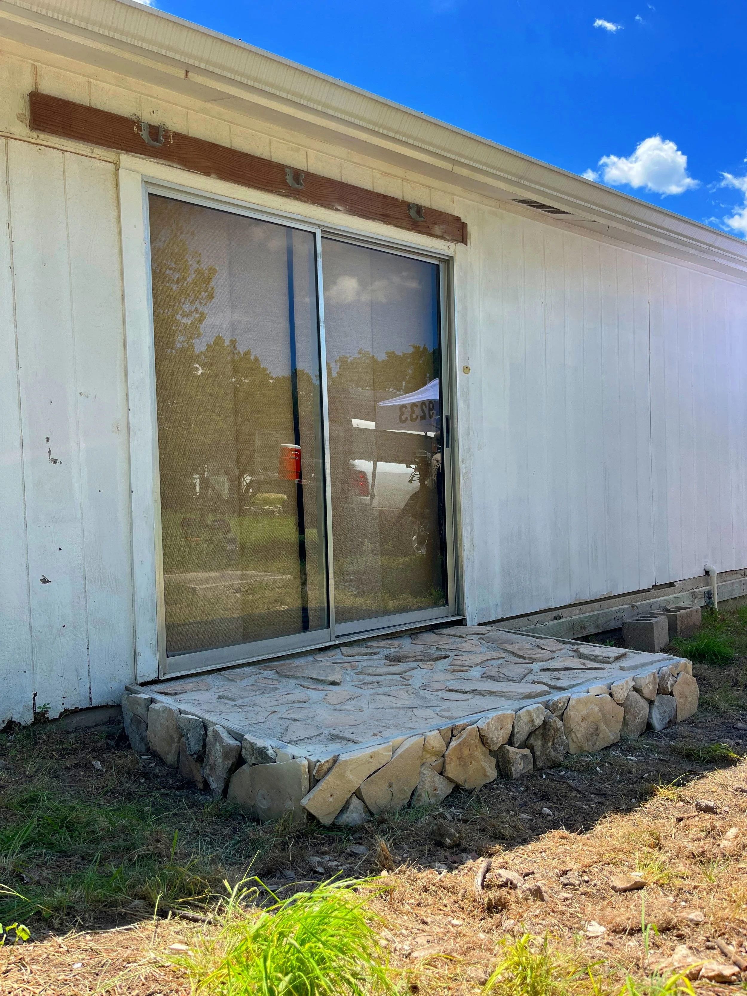 Sliding glass door installed in a white house, with a stone step leading outside. The house exterior is partly unfinished, with some wood framing above the door. The sky is clear with a few clouds.