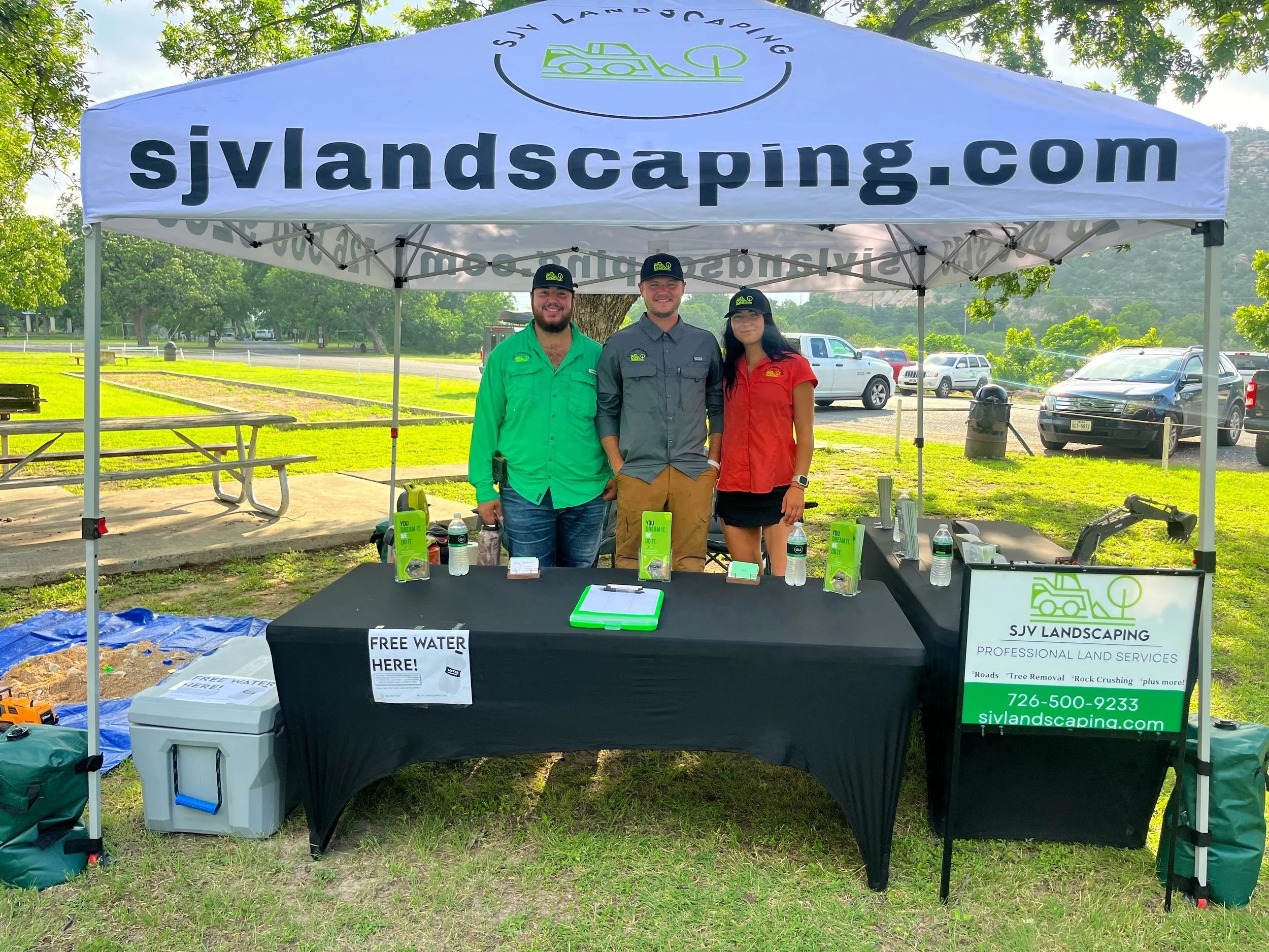 Three people standing behind a table under a canopy tent with the sign 'sjvlandscaping.com'. The table has water bottles, clipboards, and promotional materials. The tent has a logo with a tree and a vehicle, and a sign offering free water. They are o