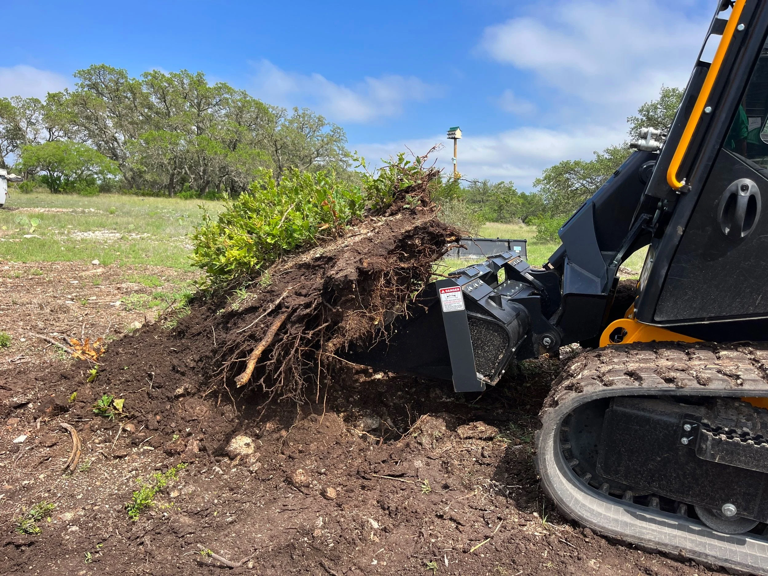 A small excavator removing a tree stump from the ground in a grassy outdoor area with trees and a partly cloudy sky.